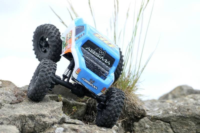 Une voiture télécommandée à la carrosserie bleue roule sur un terrain accidenté et des rochers, avec de l'herbe visible en arrière-plan.