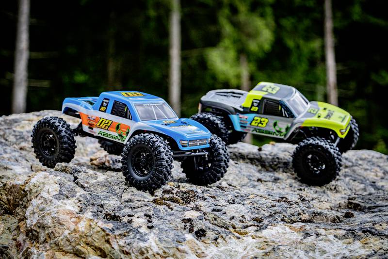 Deux voitures télécommandées roulent sur un terrain rocailleux dans la forêt. Une voiture est bleue, l'autre verte. Toutes deux ont de grandes roues.