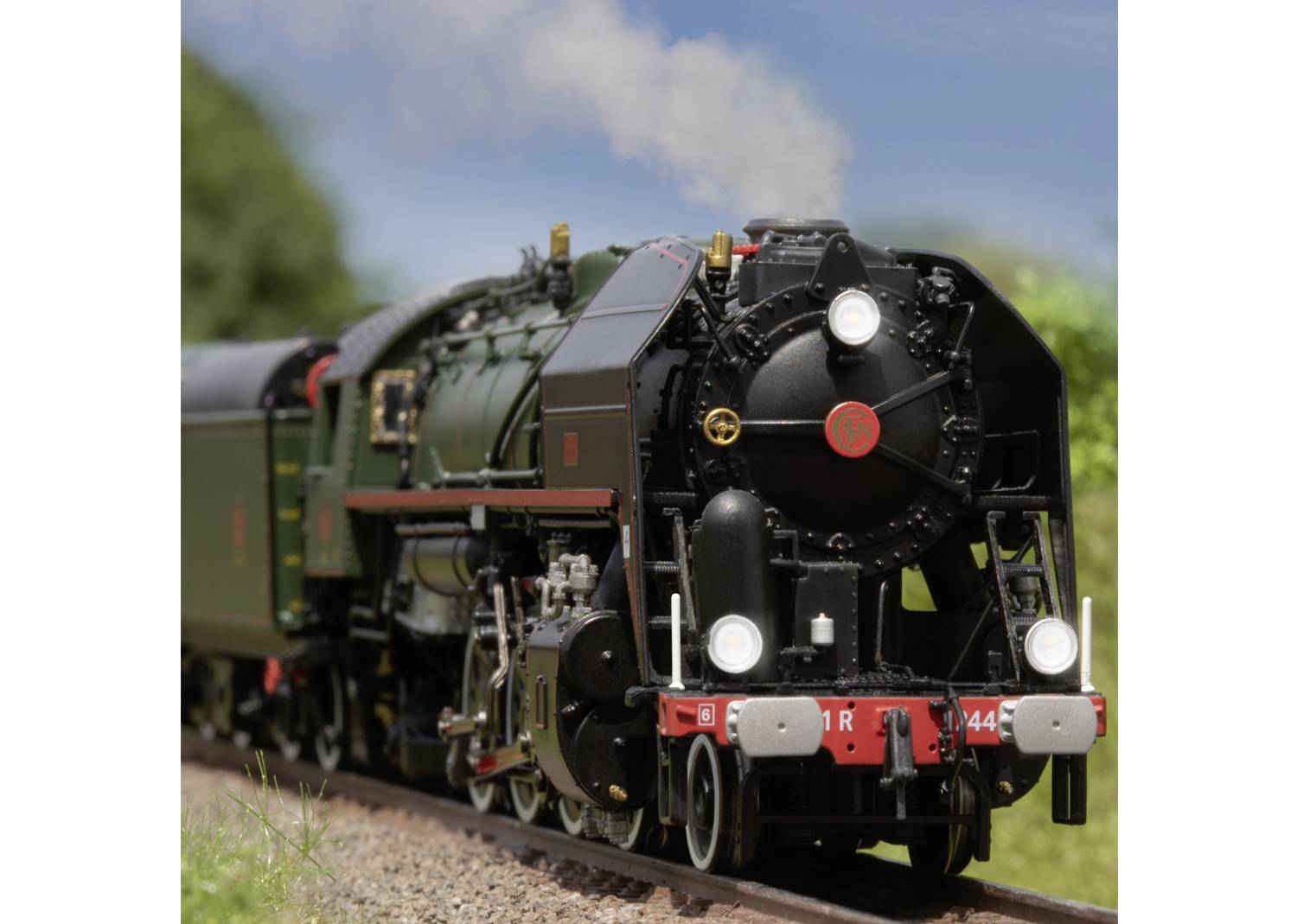 Une ancienne locomotive à vapeur sur une voie ferrée, entourée de végétation verdoyante, sous un ciel clair. La locomotive est vert foncé et noire.