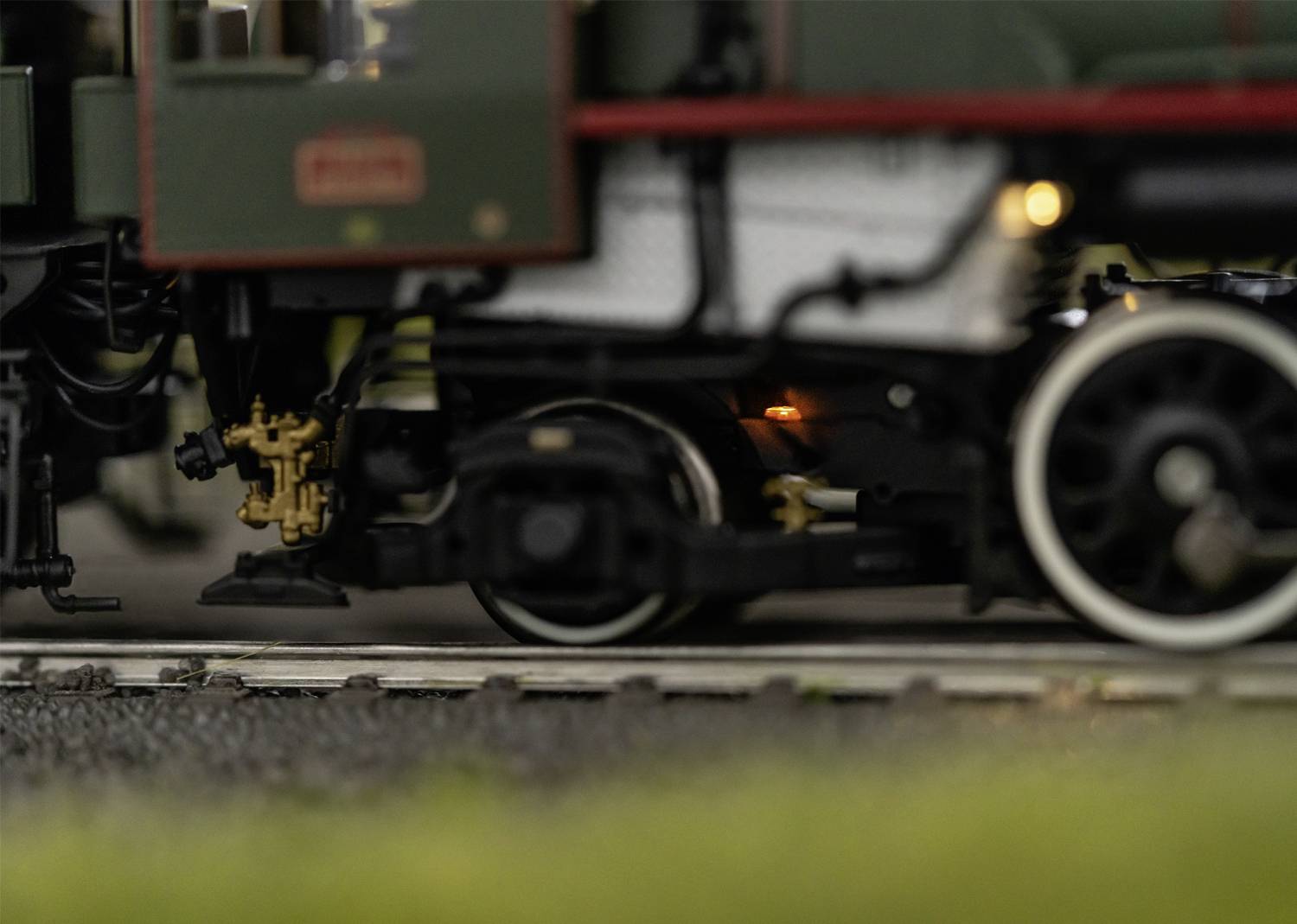 Gros plan d'un petit train miniature sur des rails. Au premier plan, des roues et des pièces mécaniques sont visibles. Arrière-plan flou.