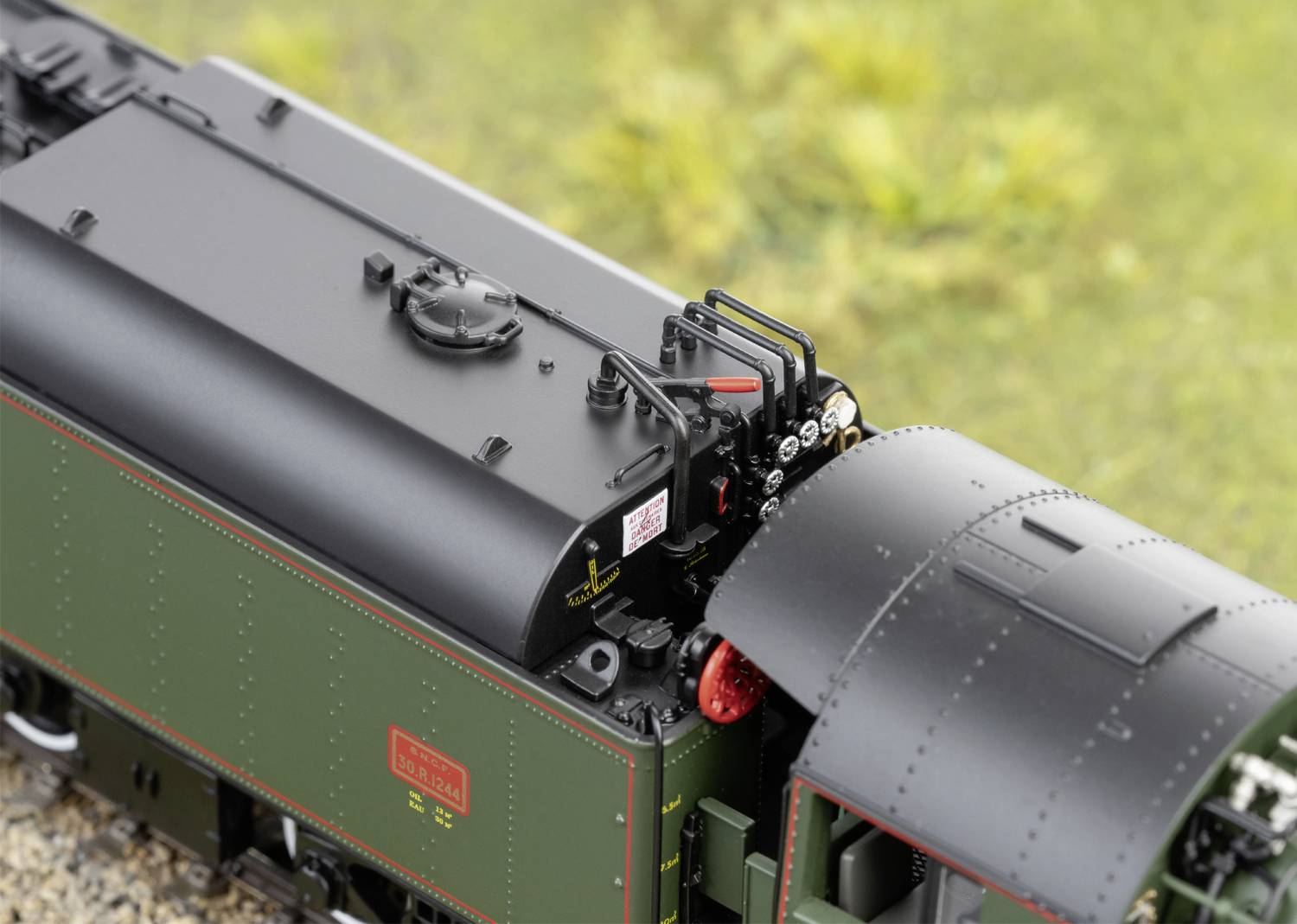 Gros plan d'une locomotive de train miniature, verte avec des accents noirs et rouges, sur des rails avec un arrière-plan flou couvert d'herbe.