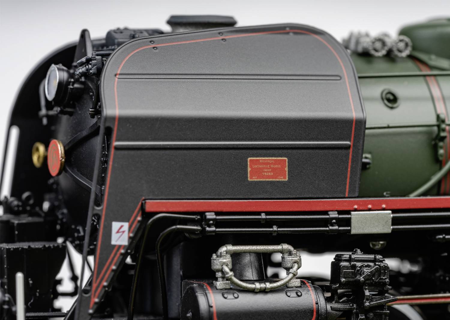 Gros plan d'une partie d'une locomotive à vapeur, révélant des détails techniques et une bande rouge sur du métal noir. Un symbole électrique est visible à gauche.
