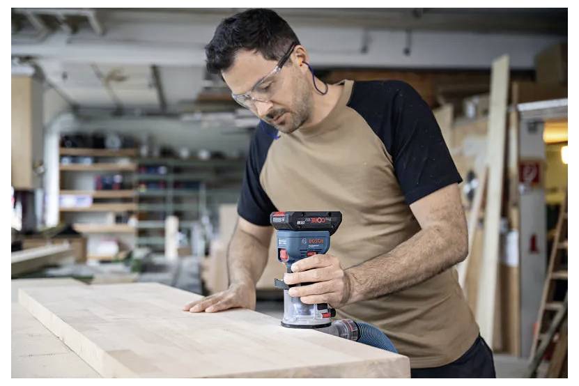 Une personne portant des lunettes de sécurité utilise une ponceuse électrique sur une surface en bois dans un atelier, avec des étagères en arrière-plan.