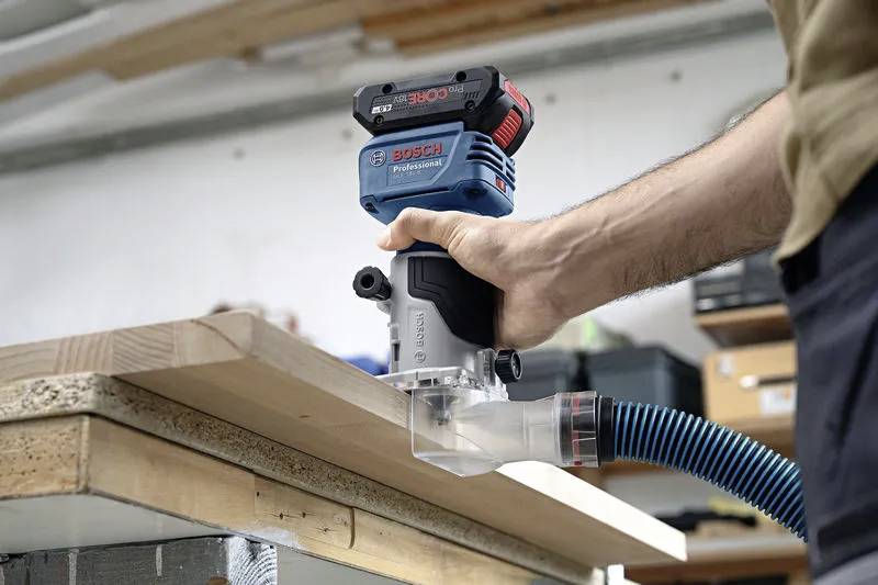 Une personne utilise une défonceuse Bosch sur une planche de bois, en se concentrant sur le travail du bois. Un dispositif de collecte de poussière est visible.