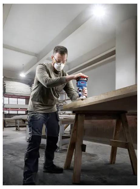Un homme masqué utilise une ponceuse électrique portative sur une table en bois dans un atelier.