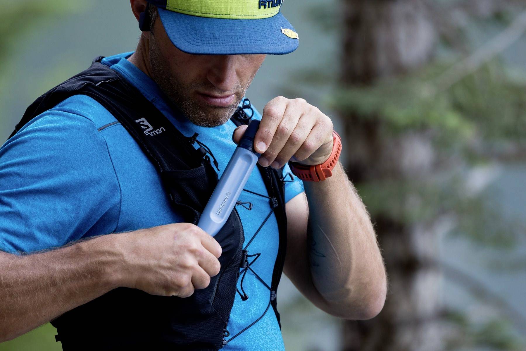Une personne remplit un sac à dos d'hydratation avec de l'eau, en extérieur dans la nature. Elle porte des vêtements bleus, une casquette et une montre orange.