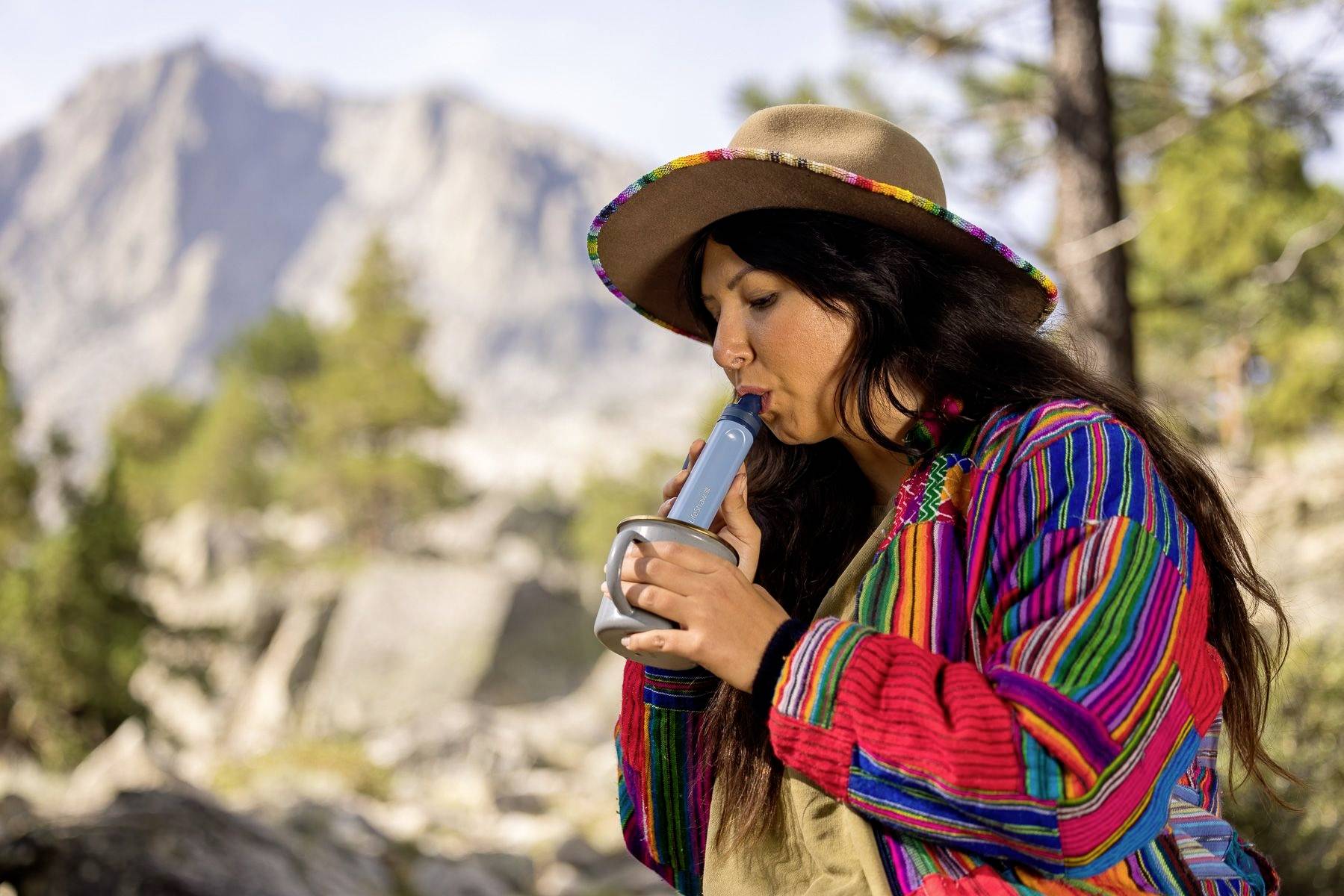 Une personne portant une veste colorée et un chapeau joue d'une flûte bleue en plein air, devant un paysage de montagne.