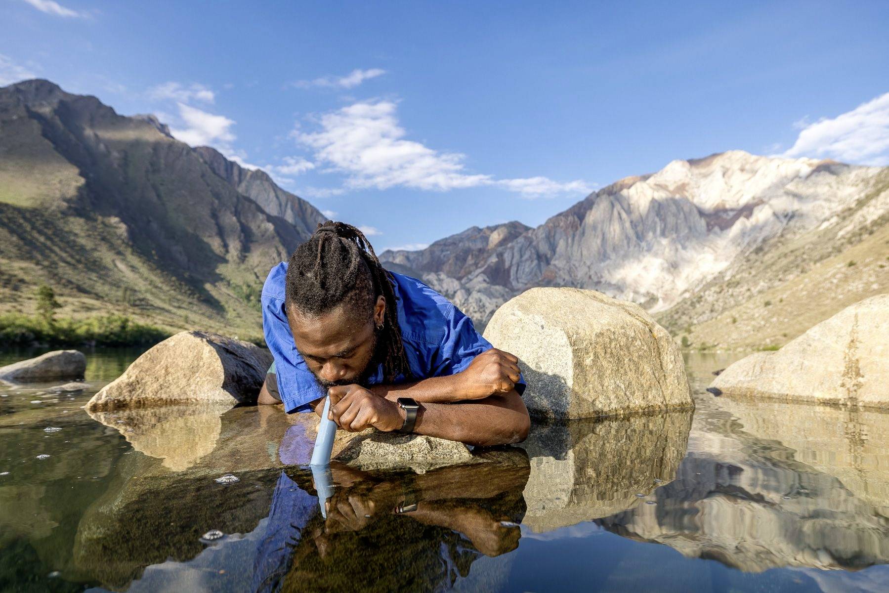 Une personne boit de l'eau dans une rivière au milieu d'un paysage montagneux sous un ciel clair.
