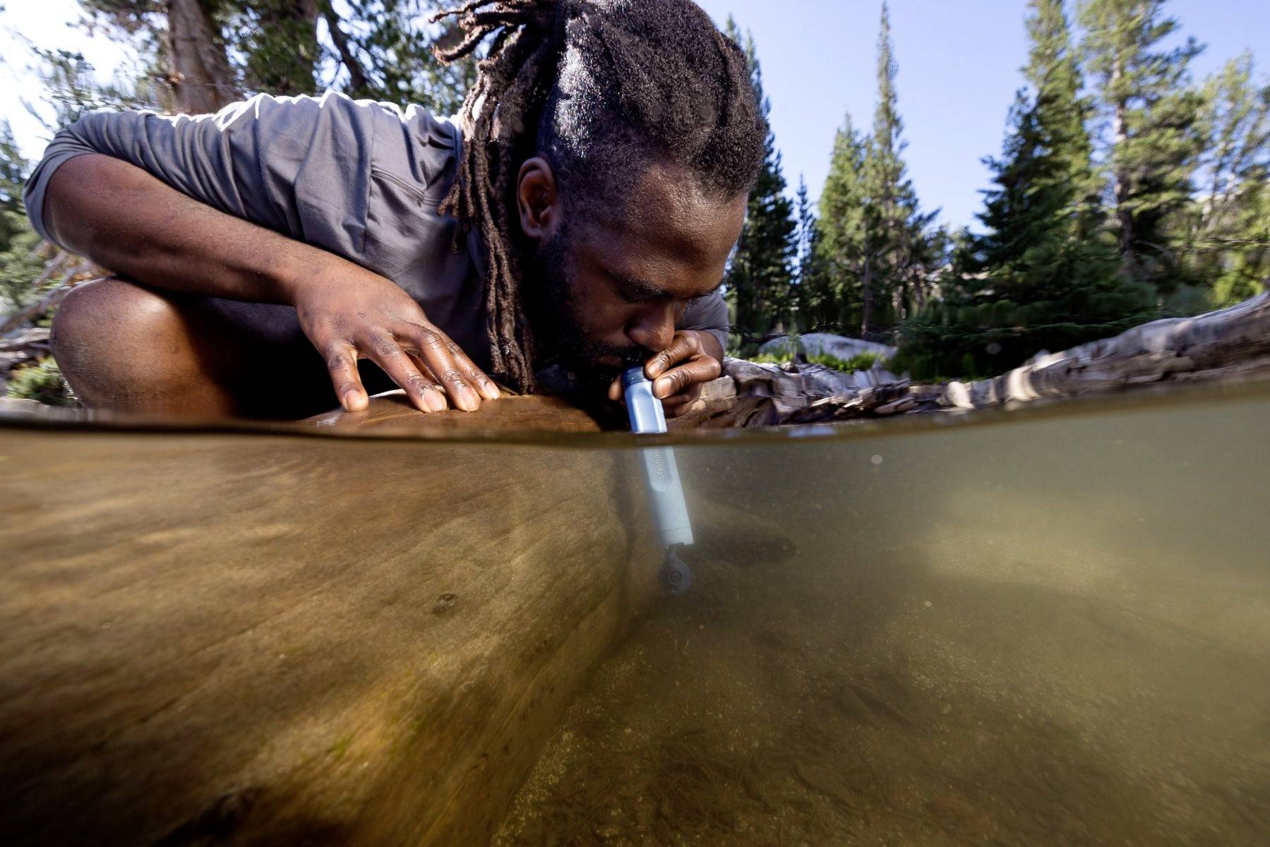 Une personne boit de l'eau à travers un filtre à eau dans un lac, entourée d'arbres et d'un ciel clair, afin d'obtenir de l'eau potable.