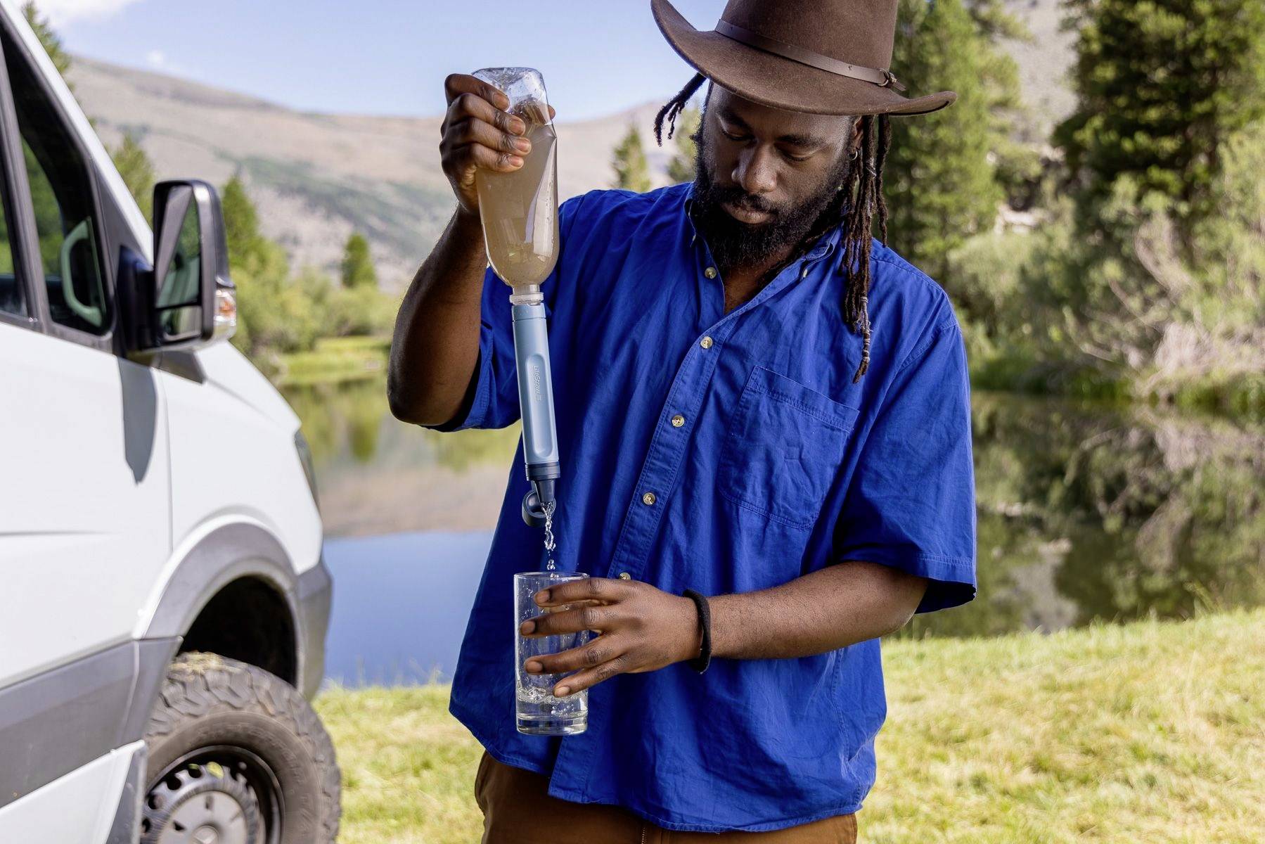 Un homme portant une chemise bleue et un chapeau filtre de l'eau dans un verre à l'aide d'un filtre à eau portable. À l'arrière-plan, on aperçoit un van, un lac et des arbres.