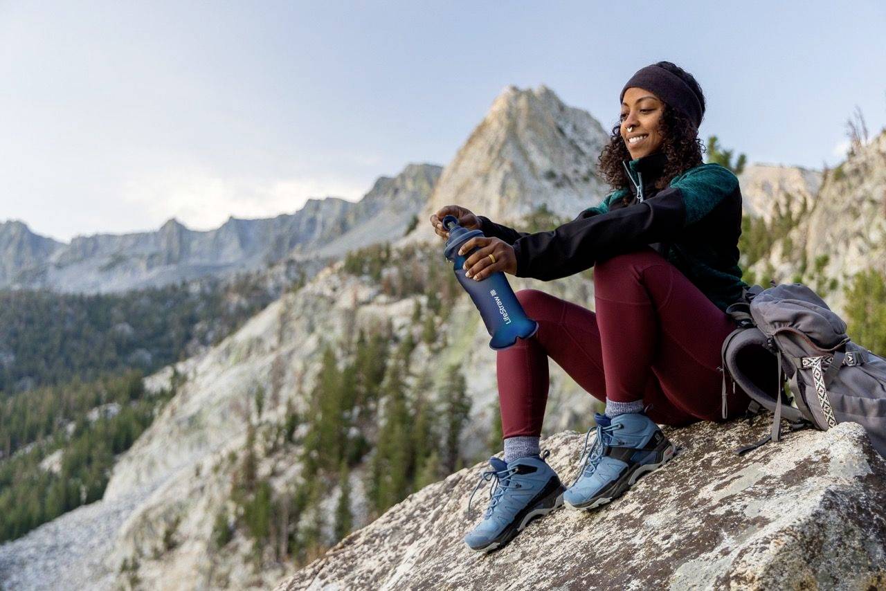 Une personne est assise sur un rocher, profitant de la vue sur les montagnes en arrière-plan et tenant une bouteille d'eau. Elle semble détendue et satisfaite.