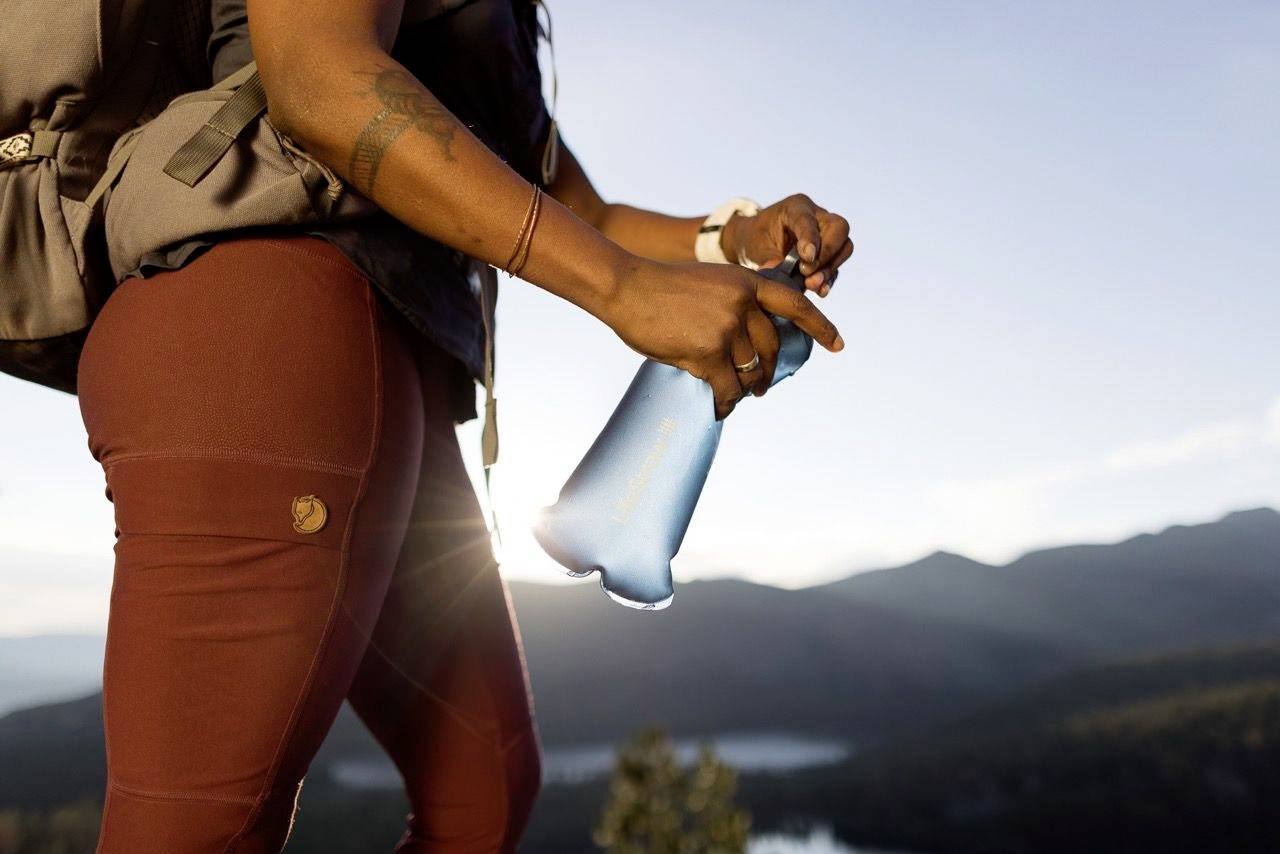 Une personne en tenue de randonnée ouvre une bouteille d'eau sur une colline avec une vue panoramique sur les montagnes en arrière-plan.