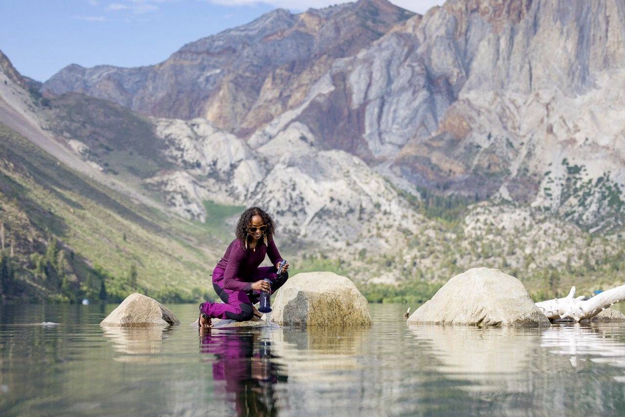 Une personne, vêtue de violet, est accroupie sur un rocher dans l'eau, devant un paysage de montagnes. Le ciel est dégagé et l'atmosphère est paisible.