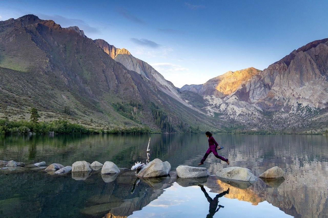 Une personne saute de pierre en pierre dans un lac de montagne calme, entouré de montagnes ensoleillées et d'un ciel bleu limpide.