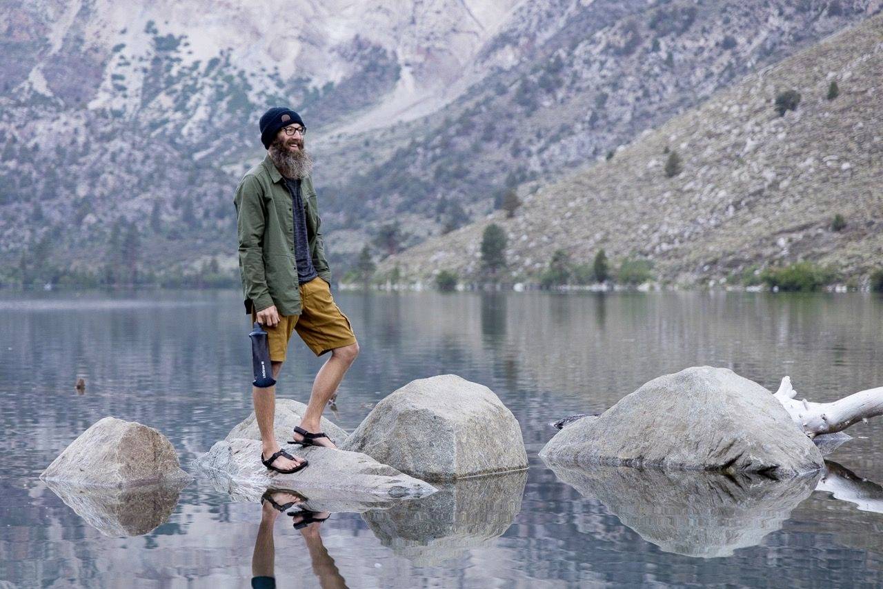 Un homme avec une barbe et une casquette se tient sur un rocher dans un lac, entouré de montagnes et d'eau réfléchissante, dans un cadre naturel détendu.