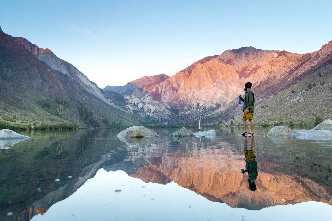 Une personne se tient sur un rocher au bord d'un lac, tenant une boisson à la main, entourée de montagnes se reflétant dans l'eau.