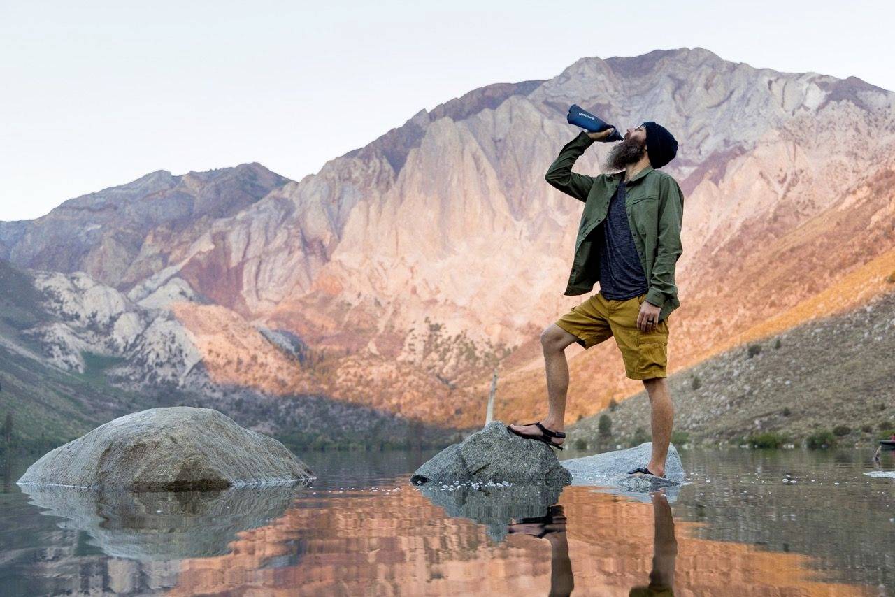 Un homme se tient sur un rocher dans l'eau et boit dans une bouteille. Des montagnes sont visibles en arrière-plan lors du lever du soleil.