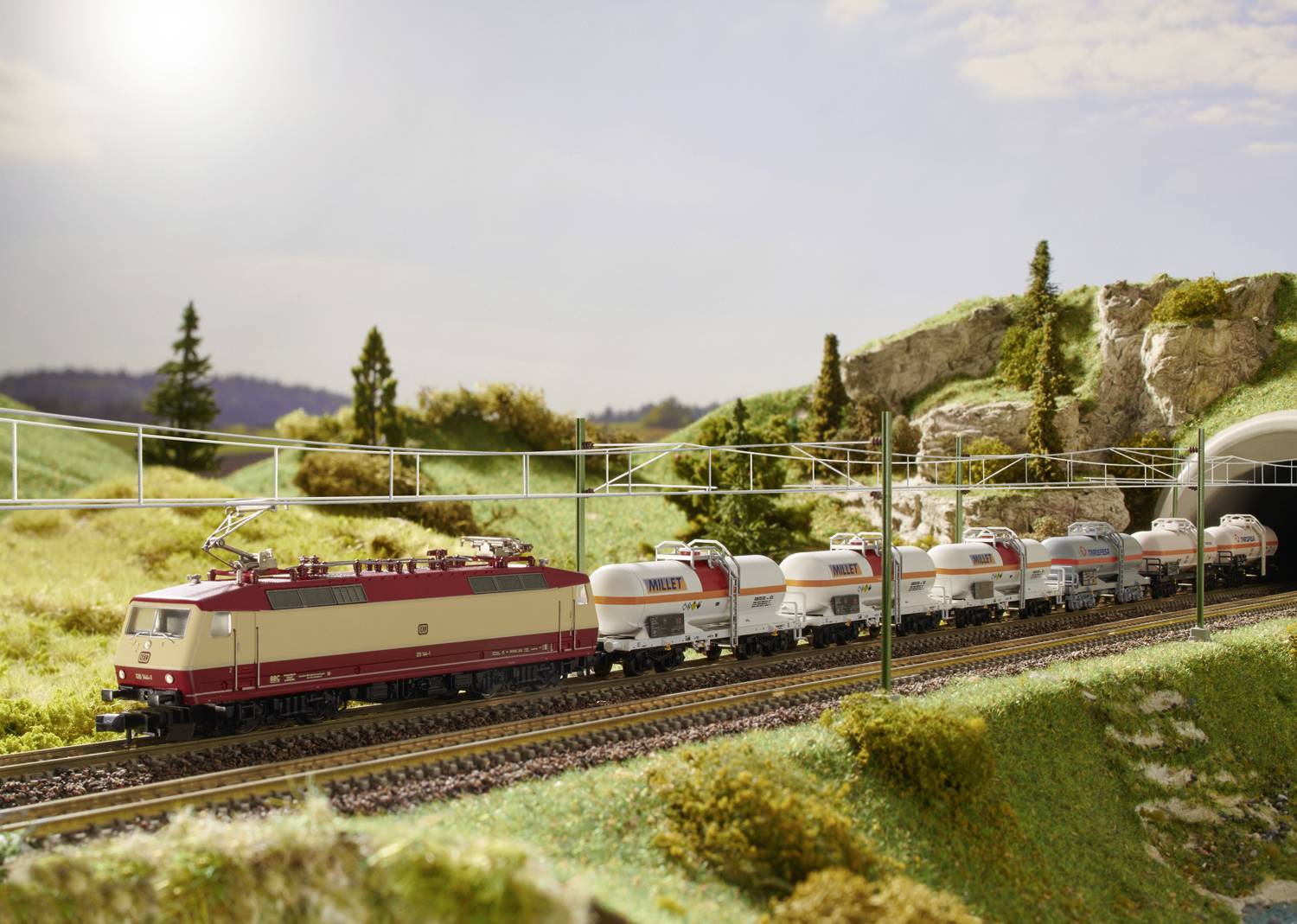 Un train miniature traverse un paysage vallonné parsemé d'arbres, tractant plusieurs wagons-citernes et s'approchant d'un tunnel.