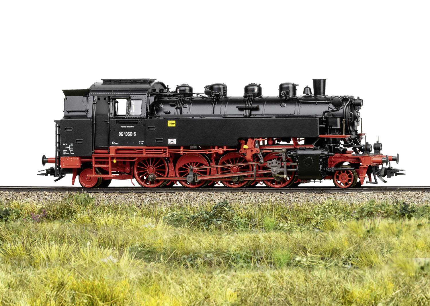 Locomotive à vapeur sur une voie ferrée, vue de profil. Chaudière noire avec châssis rouge. Entourée d'herbe.