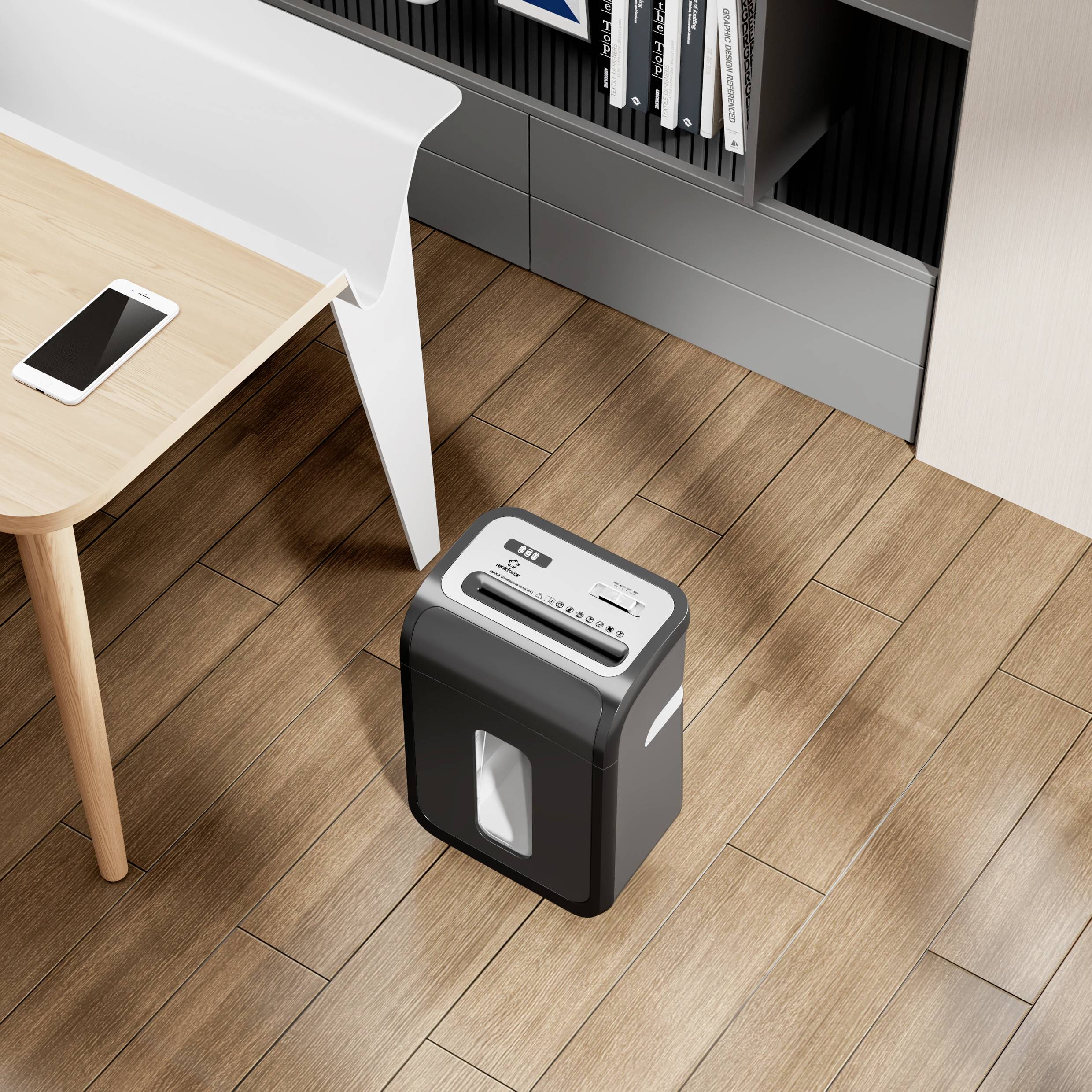Un destructeur de documents se trouve sur un plancher en bois à côté d'un bureau sur lequel repose un smartphone. Des étagères sont visibles en arrière-plan.