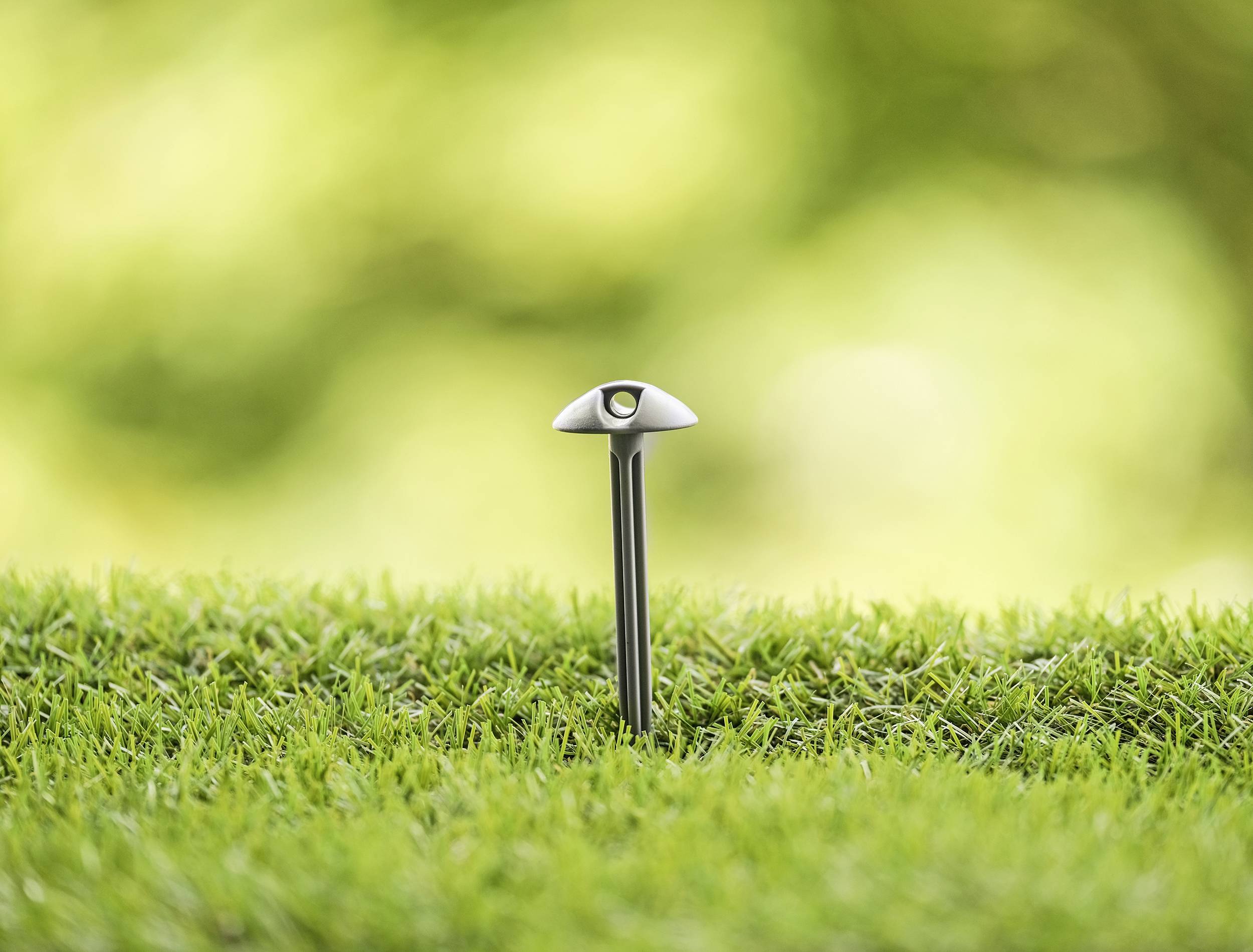 Un poteau métallique moderne et isolé, équipé d'un petit élément lumineux, se dresse dans une pelouse dense et verte devant un arrière-plan flou.