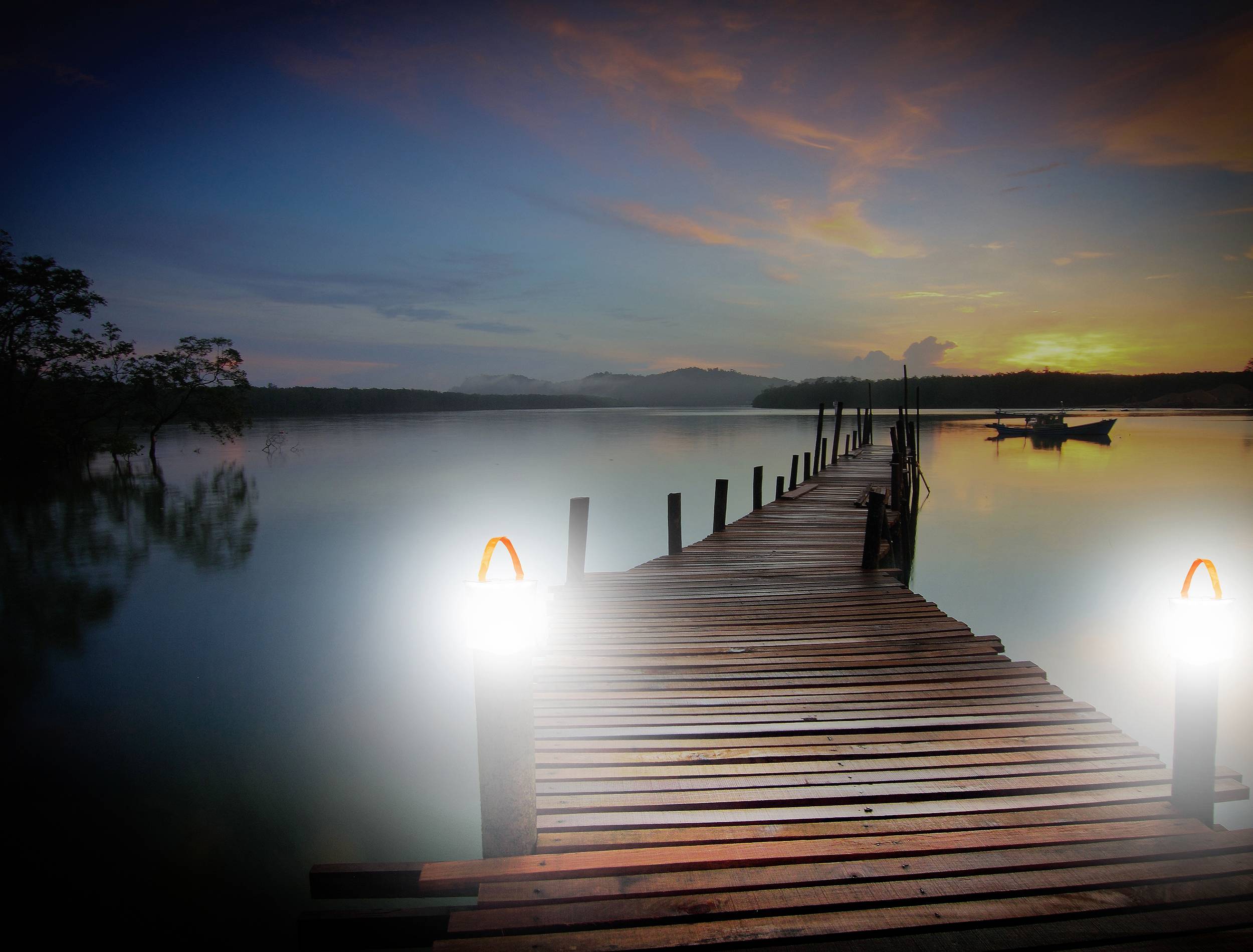 Une longue passerelle en bois bordée de lanternes mène à un lac paisible au coucher du soleil. Un petit bateau est visible en arrière-plan.