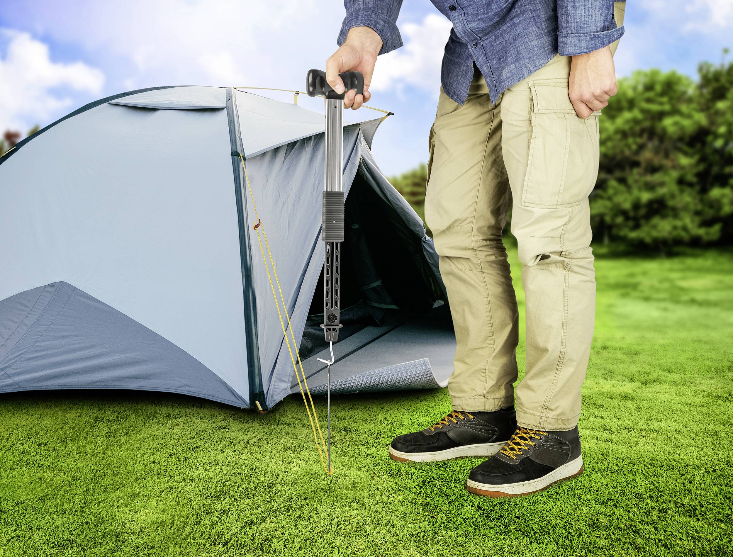 Une personne tend une tente à l'aide d'un outil de tension sur l'herbe verte. La tente est bleue et le ciel est dégagé.
