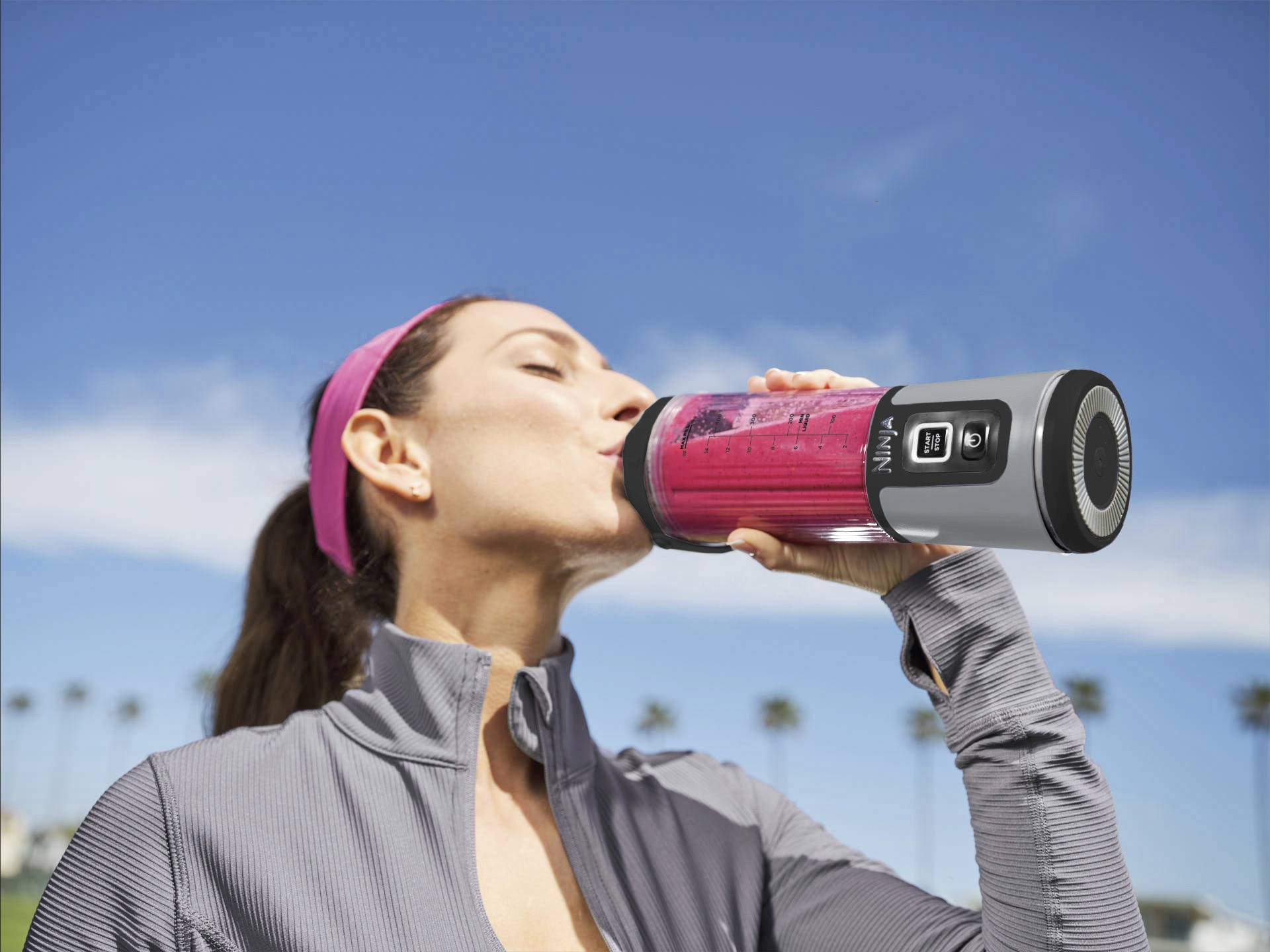 Une femme boit dans un mixeur portable contenant une boisson rouge, avec un ciel bleu en arrière-plan. Elle porte un bandeau et des vêtements de sport.