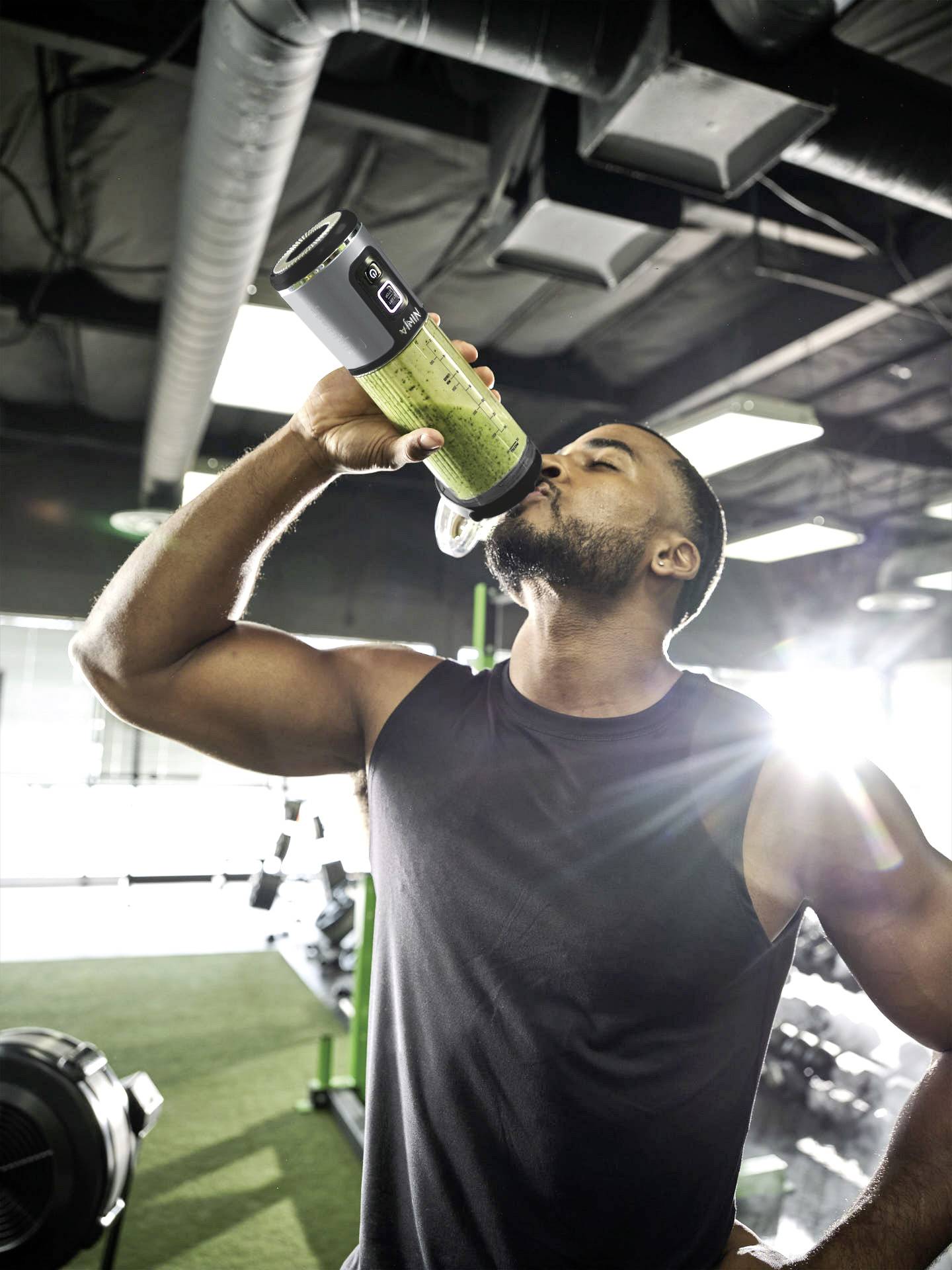 Un homme dans une salle de sport boit dans une bouteille de sport. Des équipements de fitness sont visibles en arrière-plan. La lumière du soleil pénètre à travers les fenêtres.