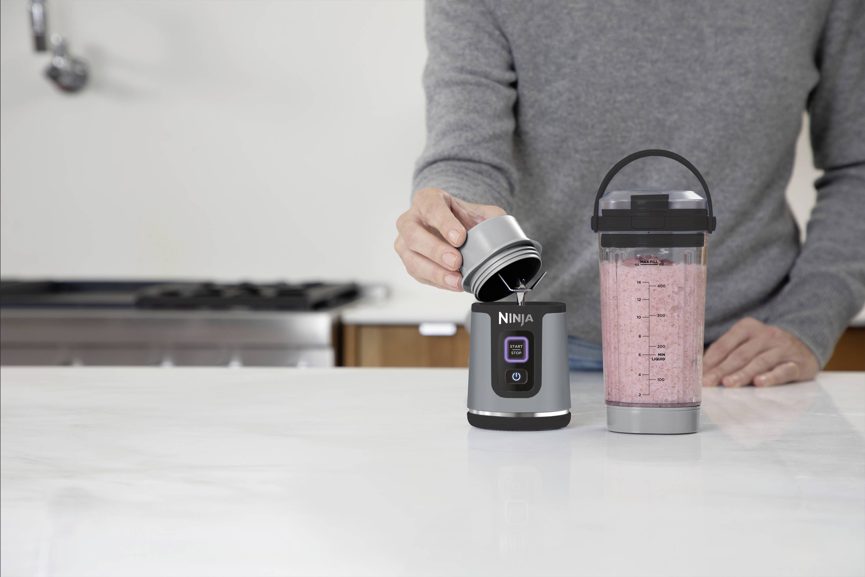 Une personne prépare des smoothies avec un mixeur sur socle. Le mixeur est posé sur un plan de travail blanc, à côté duquel se trouve un gobelet rempli.