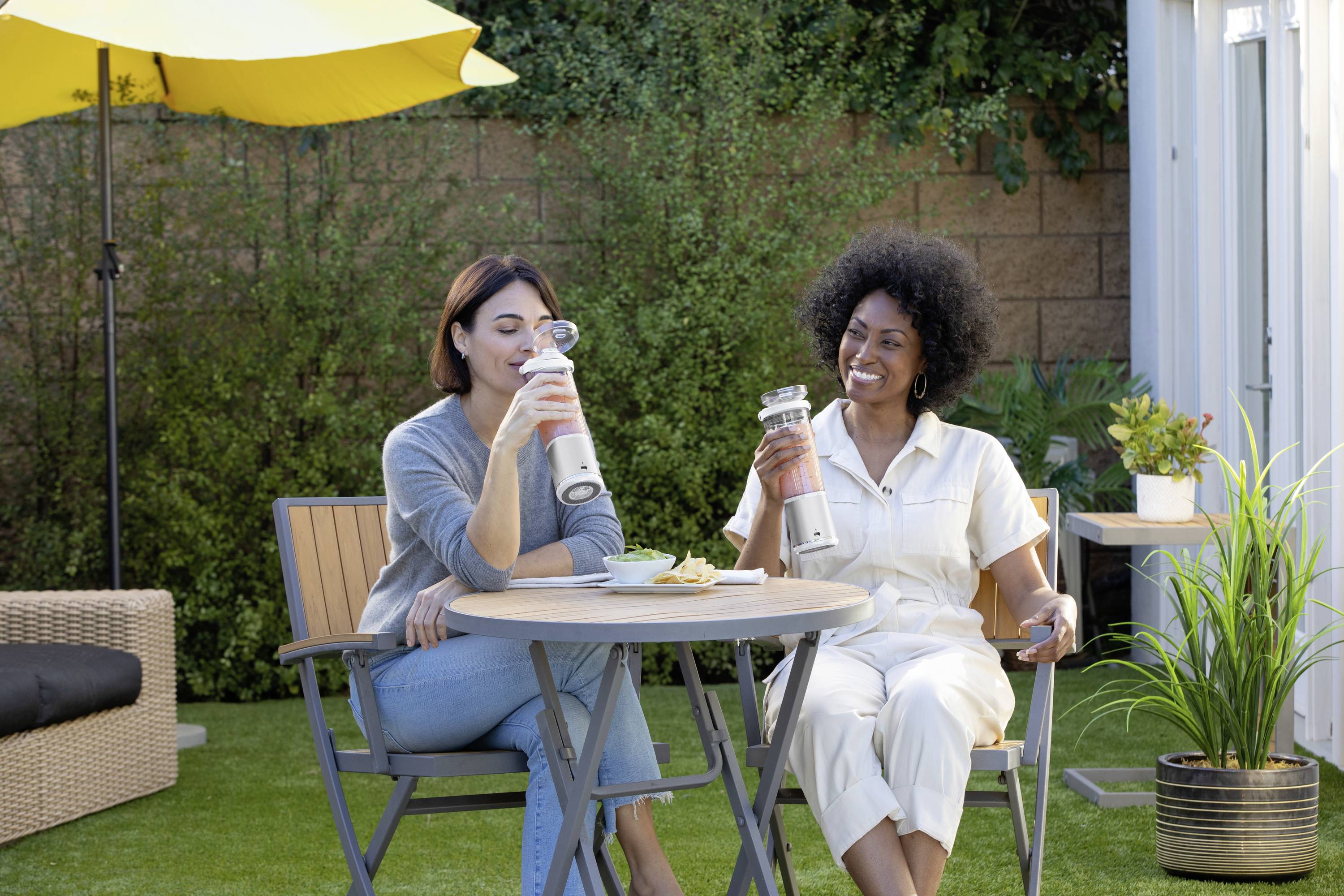 Deux femmes sont assises à une table dans un jardin et boivent dans des gourdes. Il fait soleil, et des plantes sont visibles en arrière-plan.