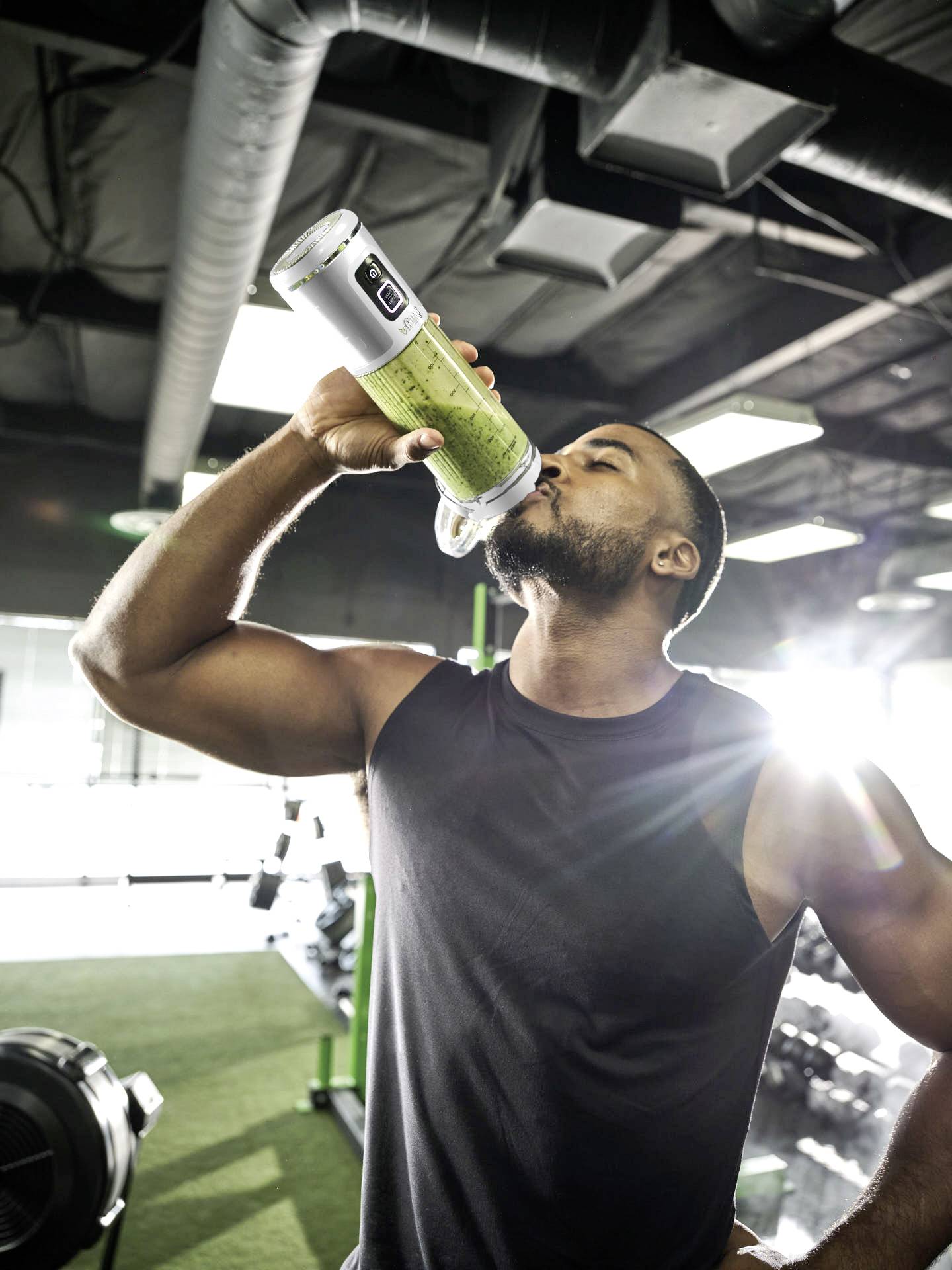 Un homme dans une salle de sport boit dans une bouteille contenant une boisson verte. La lumière du soleil brille depuis l'arrière. Des équipements de fitness sont visibles.