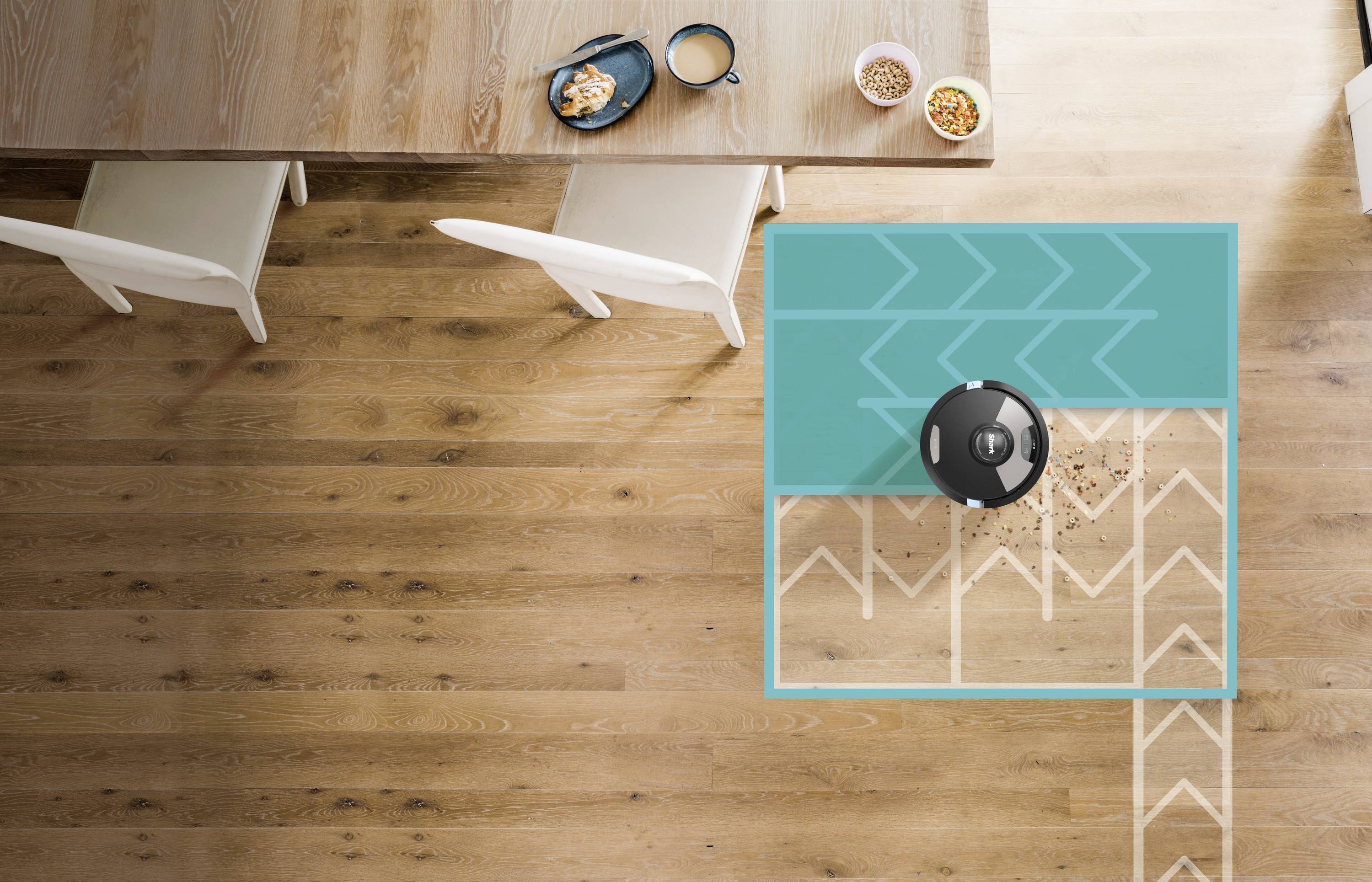 Un aspirateur robot nettoie un sol en bois à côté d'une table à manger avec deux chaises claires et de la vaisselle posée dessus.