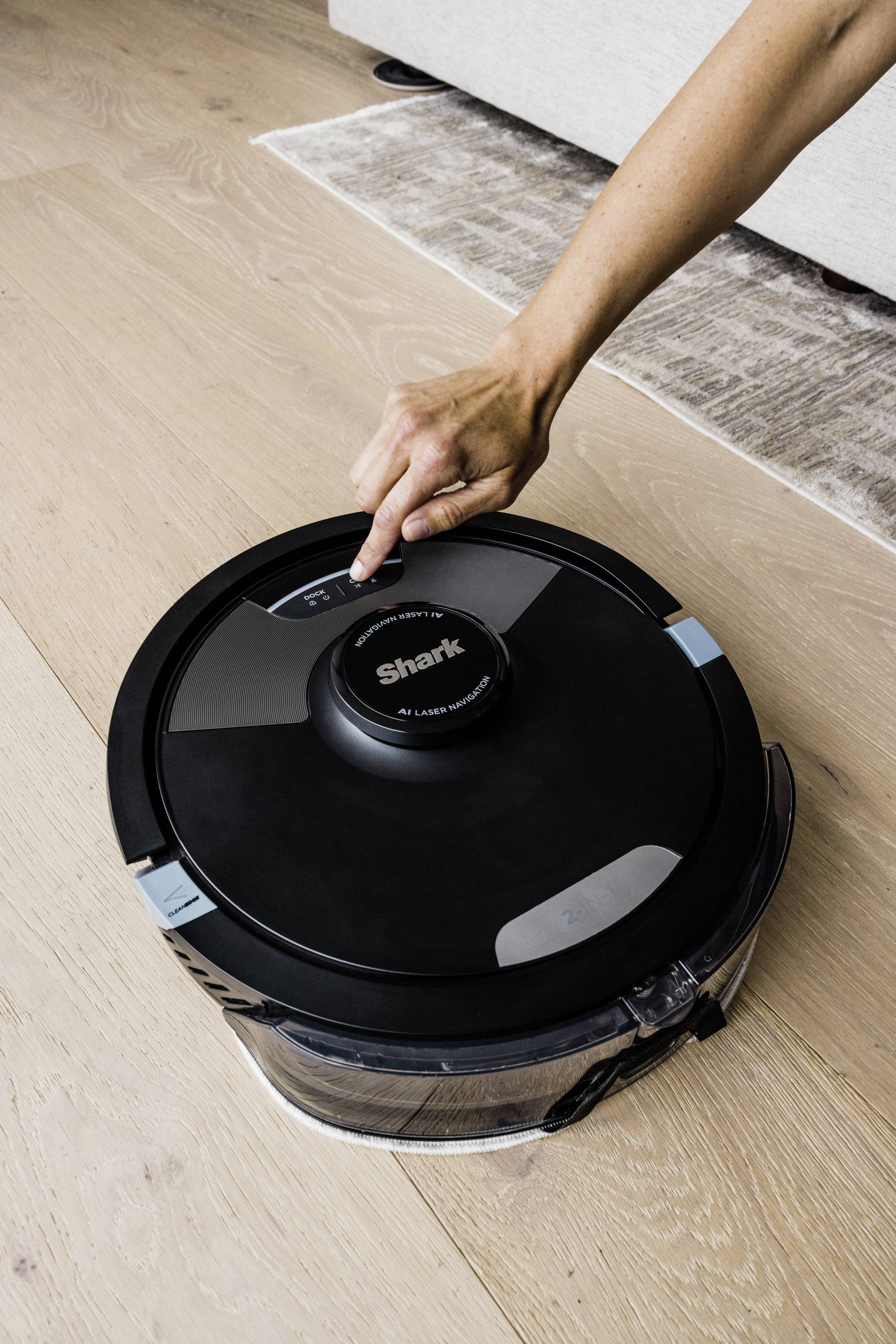 Une main appuie sur un robot aspirateur rond noir sur un sol en bois. Le robot est de la marque 