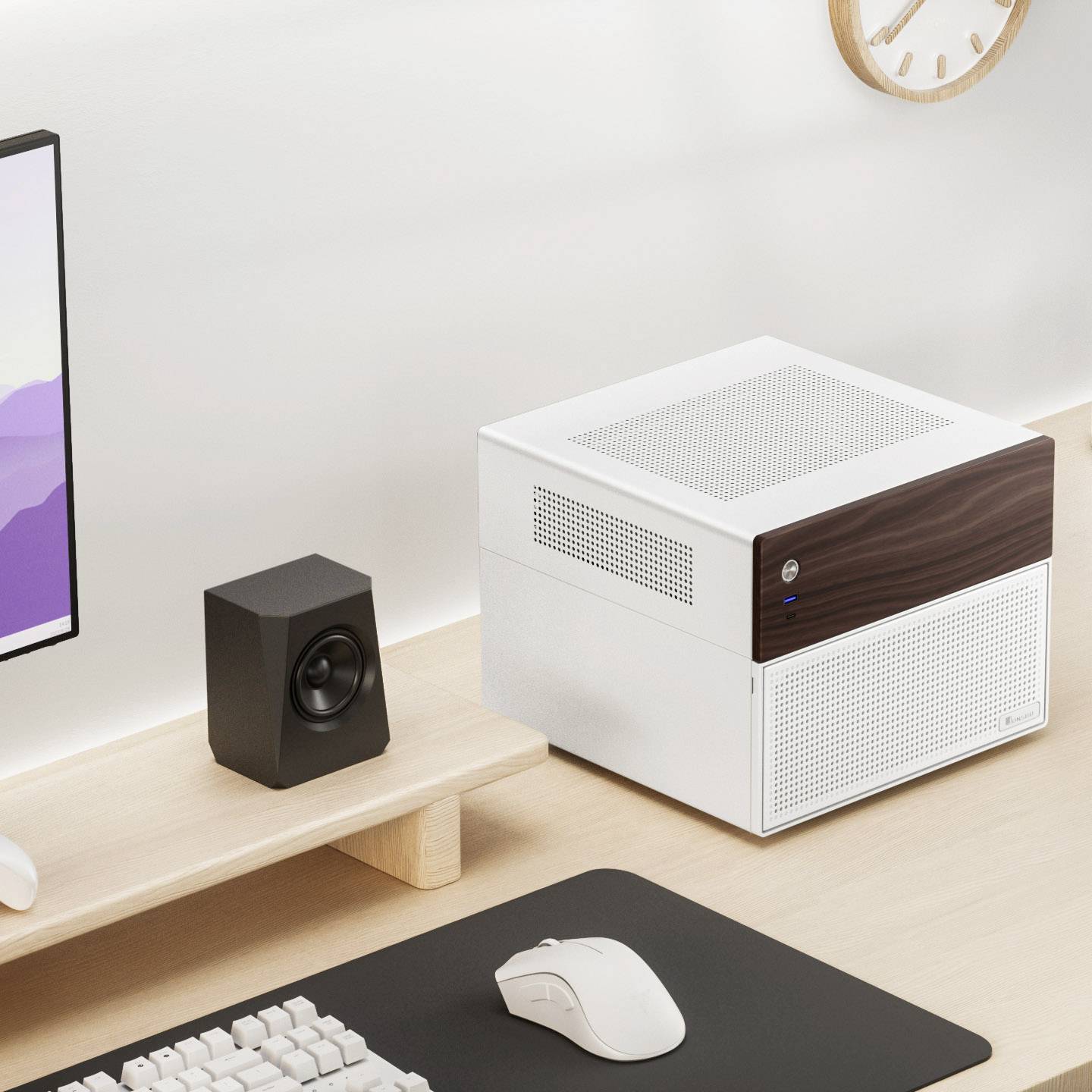 Un bureau avec un petit ordinateur blanc, à côté un haut-parleur, une souris et un clavier. Une horloge murale en arrière-plan.