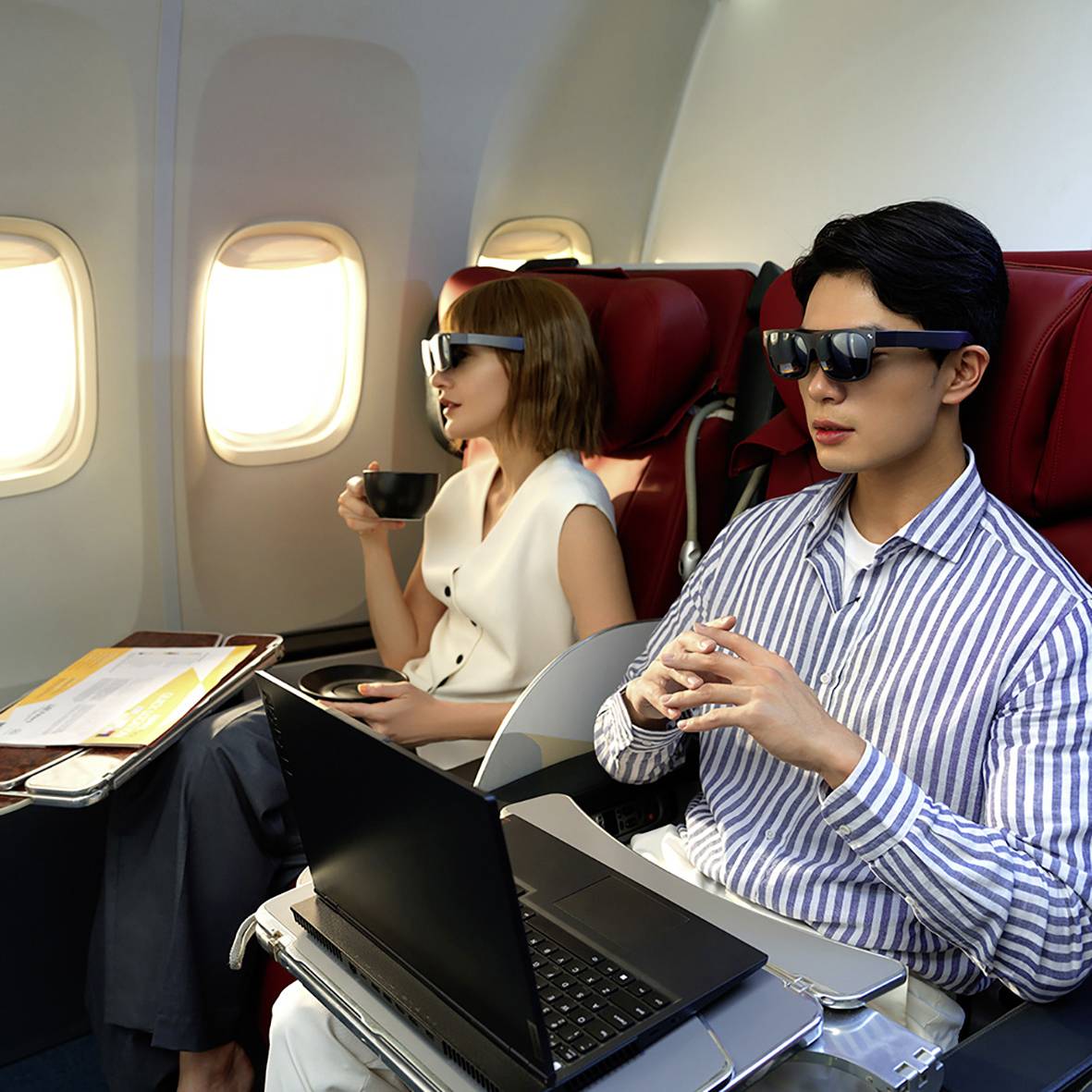 Un homme dans l'avion porte des lunettes de soleil et travaille sur un ordinateur portable. À côté de lui, une femme boit dans un gobelet en carton.