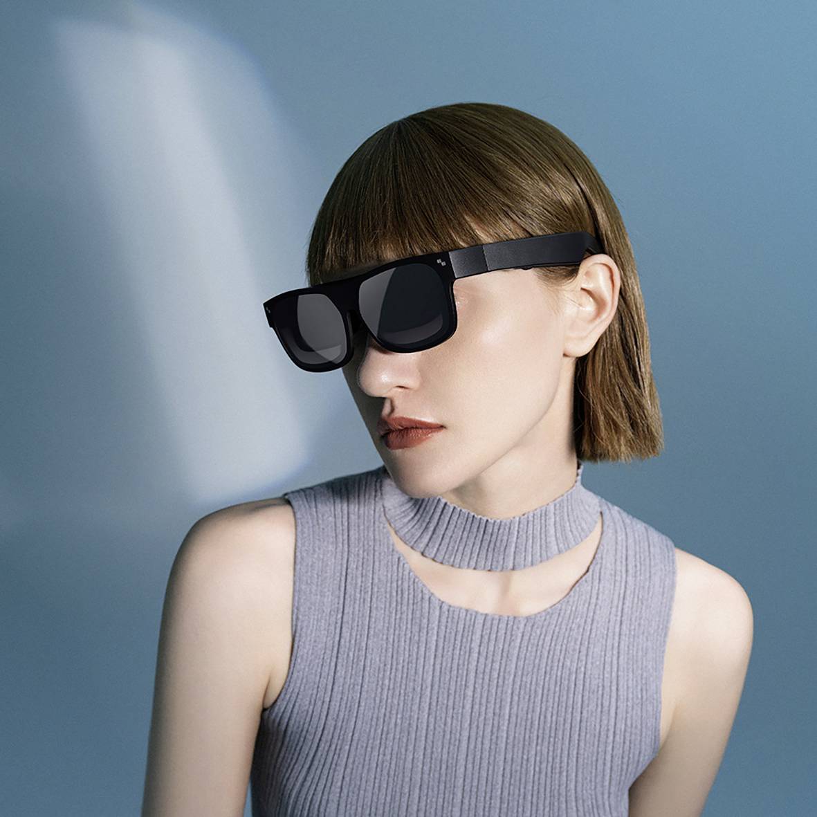 Une personne aux cheveux lisses porte des lunettes de réalité virtuelle noires futuristes et un haut gris sans manches, assise dans un espace neutre.