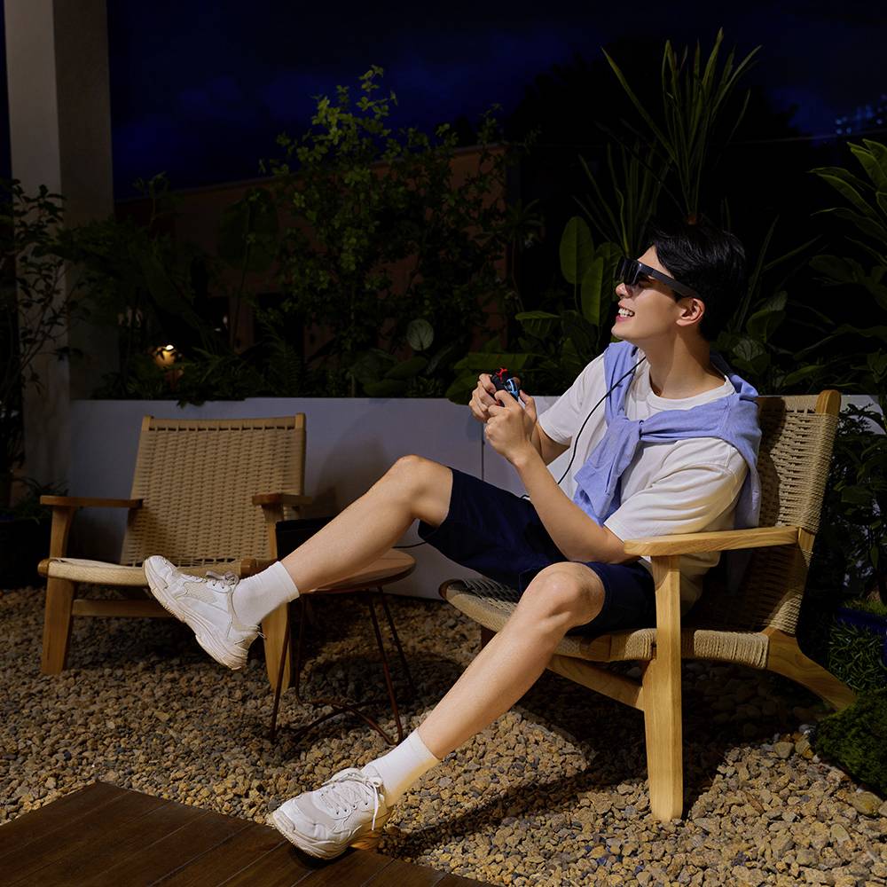 Un homme est assis dans un fauteuil de jardin la nuit, souriant, portant des lunettes de soleil sombres et tenant un petit emballage rouge.