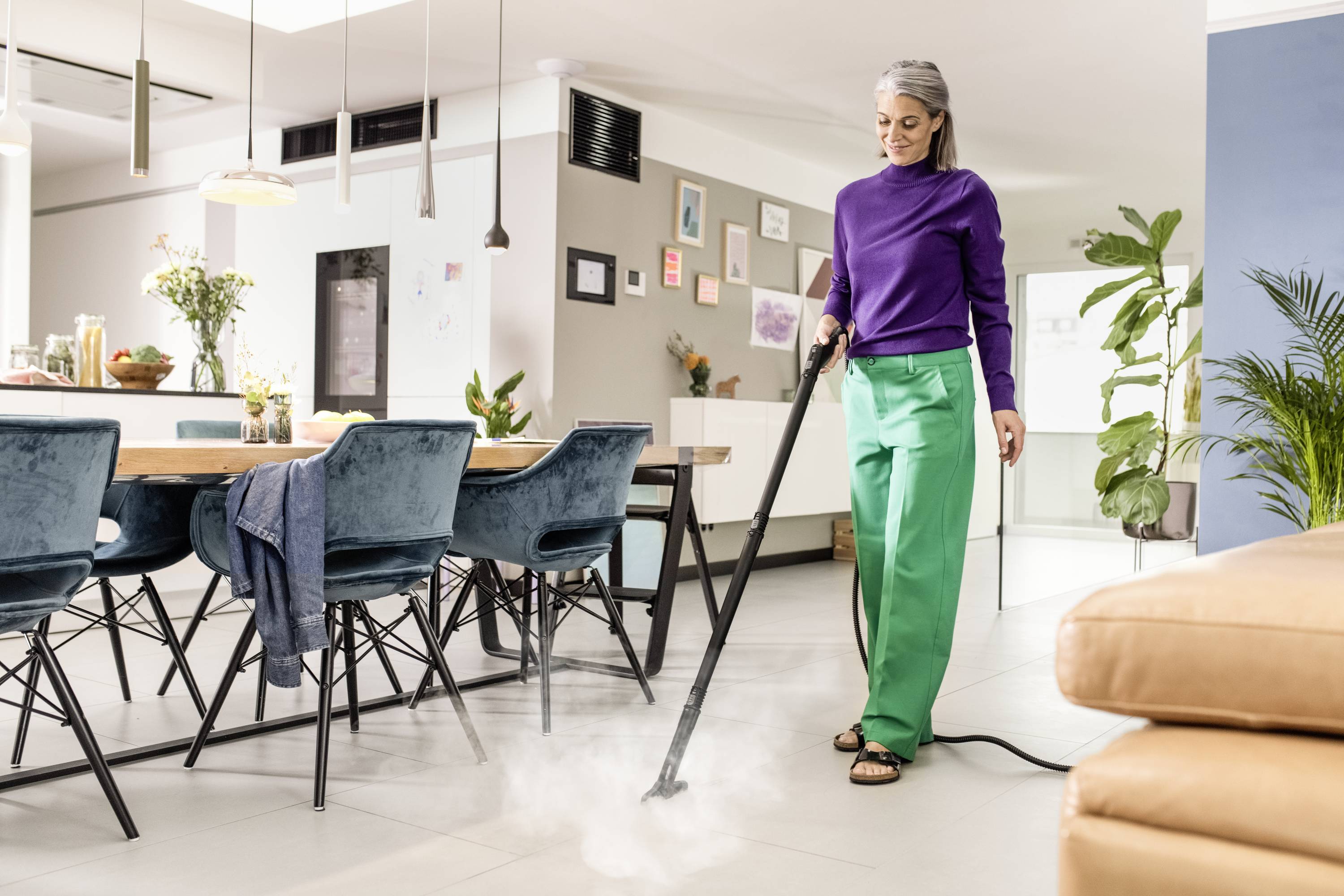 Une personne nettoie le sol avec un nettoyeur vapeur dans un salon moderne et élégamment aménagé, avec une zone repas.