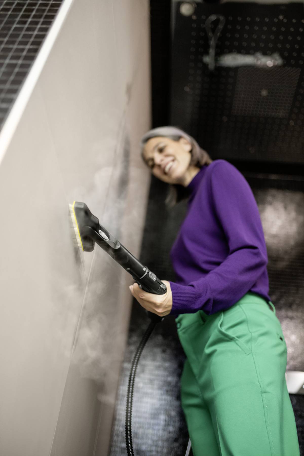 Une personne portant un pull violet et un pantalon vert utilise un nettoyeur vapeur sur un mur carrelé, avec un léger sourire.