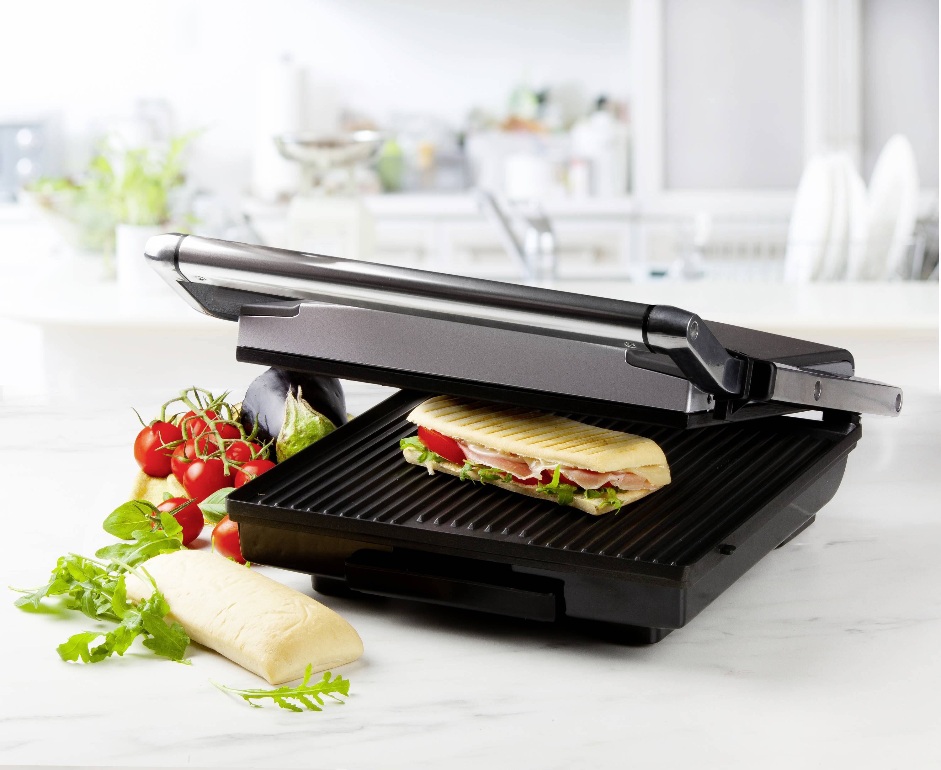 Un grill à contact ouvert avec un panini et des légumes frais à côté sur un plan de travail de cuisine.