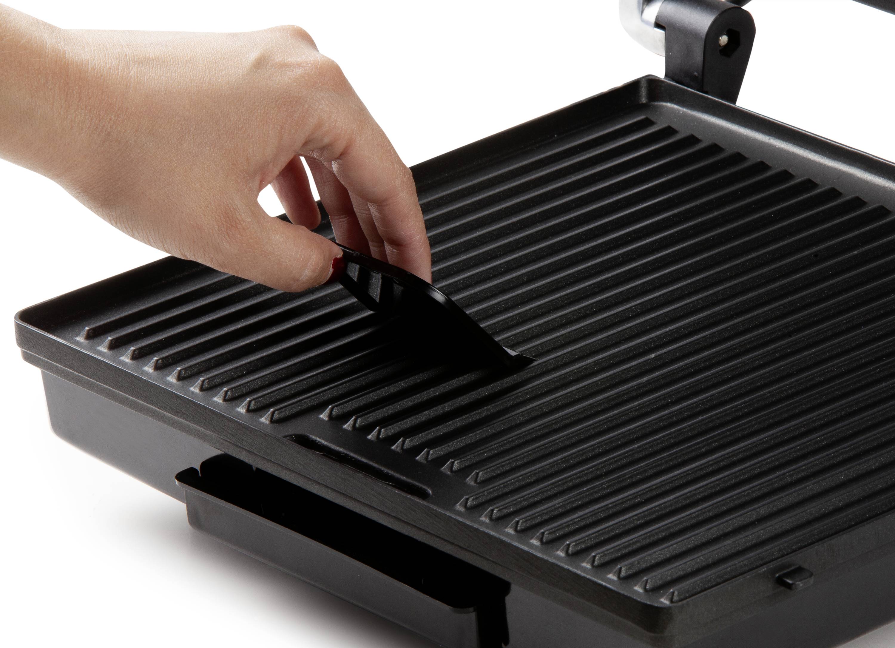 Une main soulève la grille d'un grill de contact. Le grill présente une surface striée, adaptée pour griller de la viande ou des légumes.