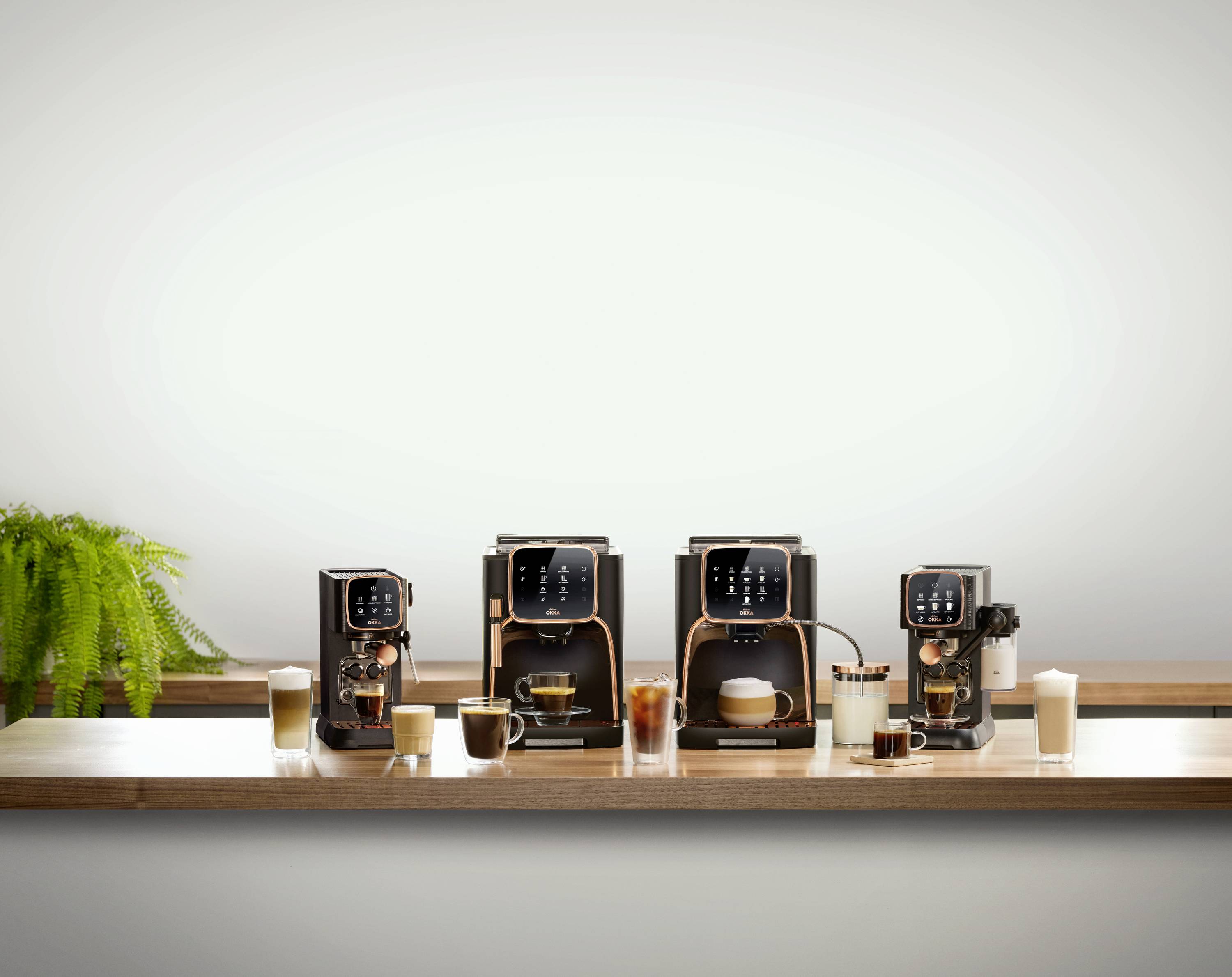 Trois machines à café modernes alignées sur une table, avec différentes boissons café préparées et une plante en pot à gauche.
