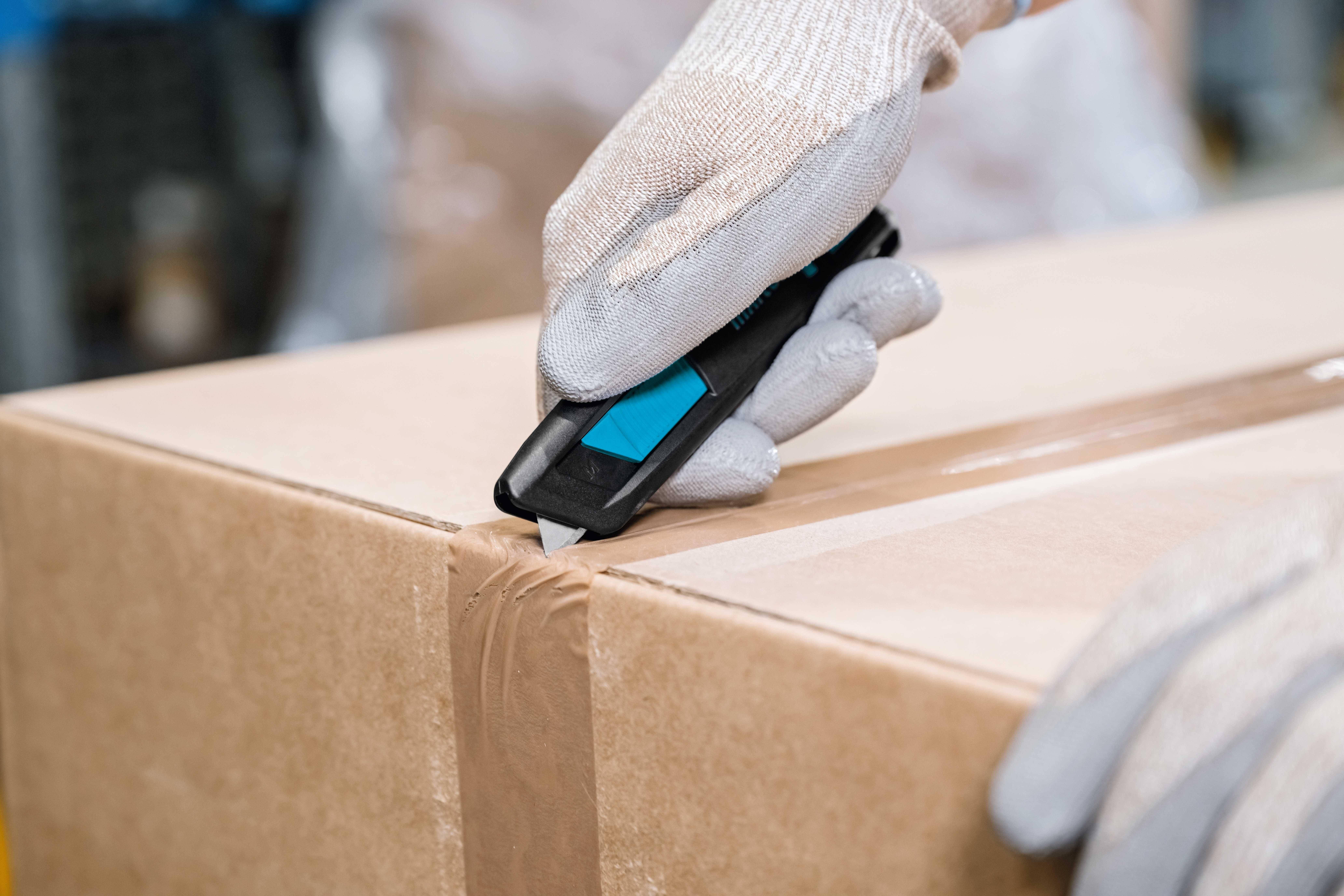 Une main gantée ouvre un carton avec un cutter.