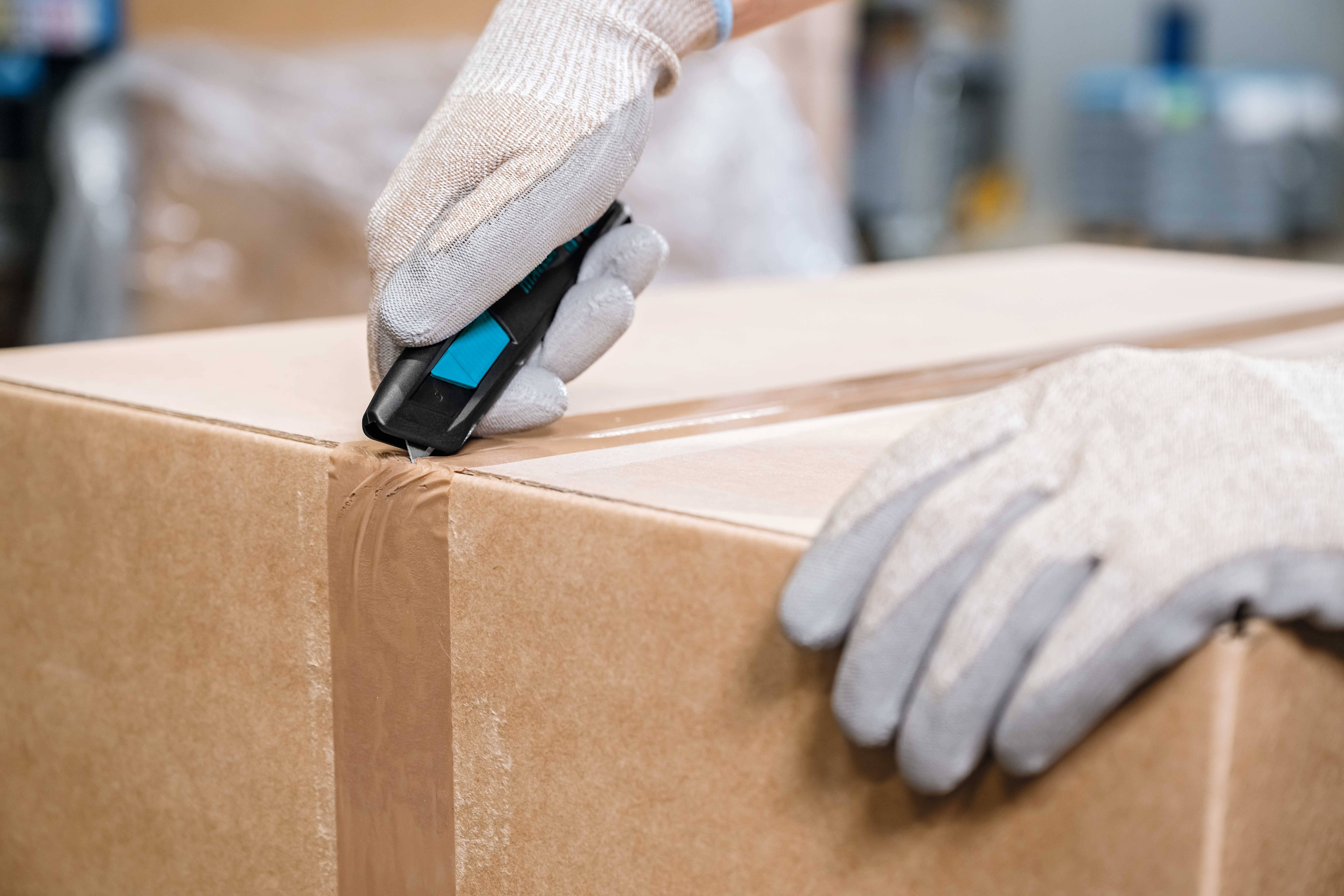 Une personne portant des gants ouvre un carton à l'aide d'un cutter.
