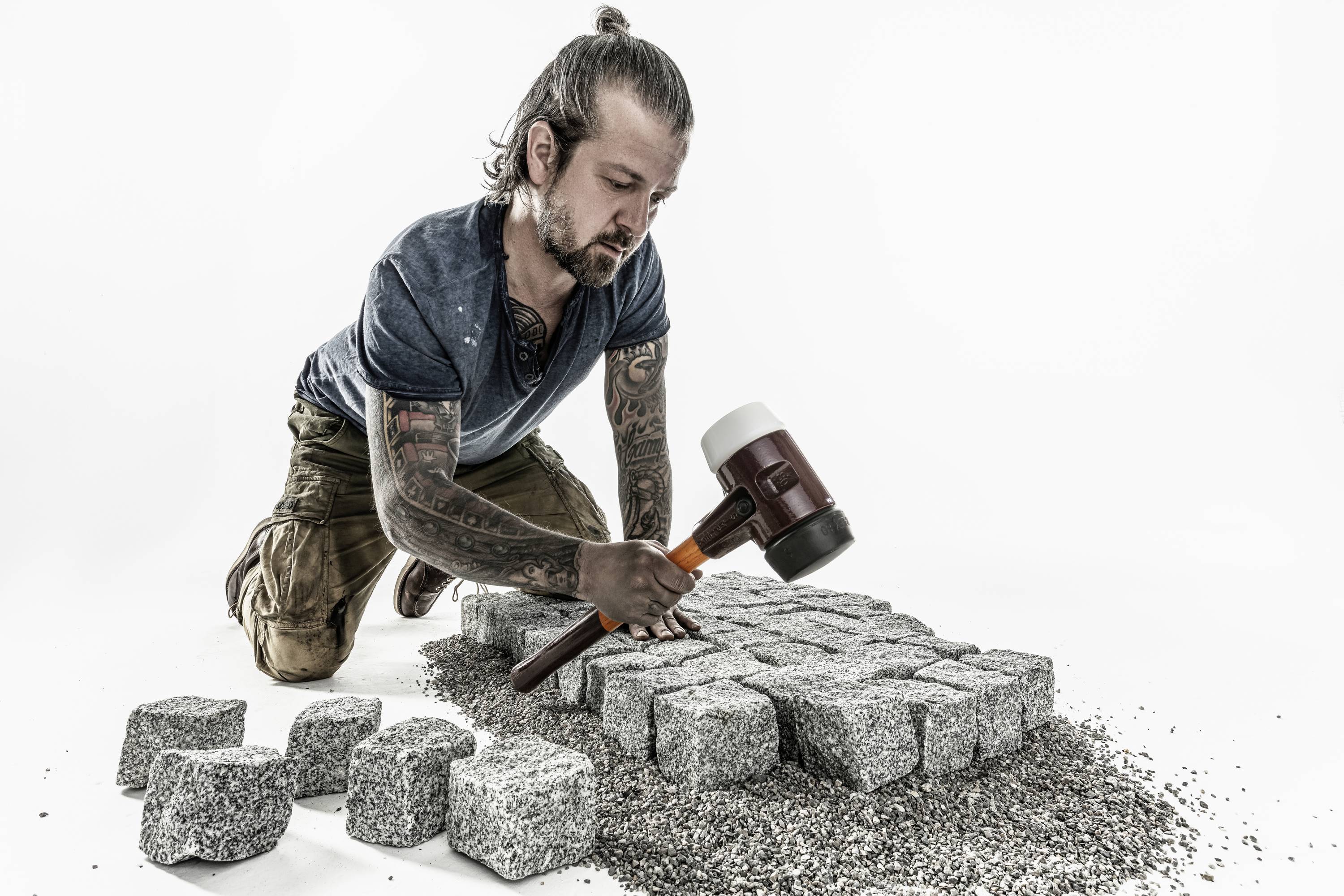 Un ouvrier est agenouillé au sol et pose des pavés avec un maillet en caoutchouc. La scène illustre la précision artisanale au travail.