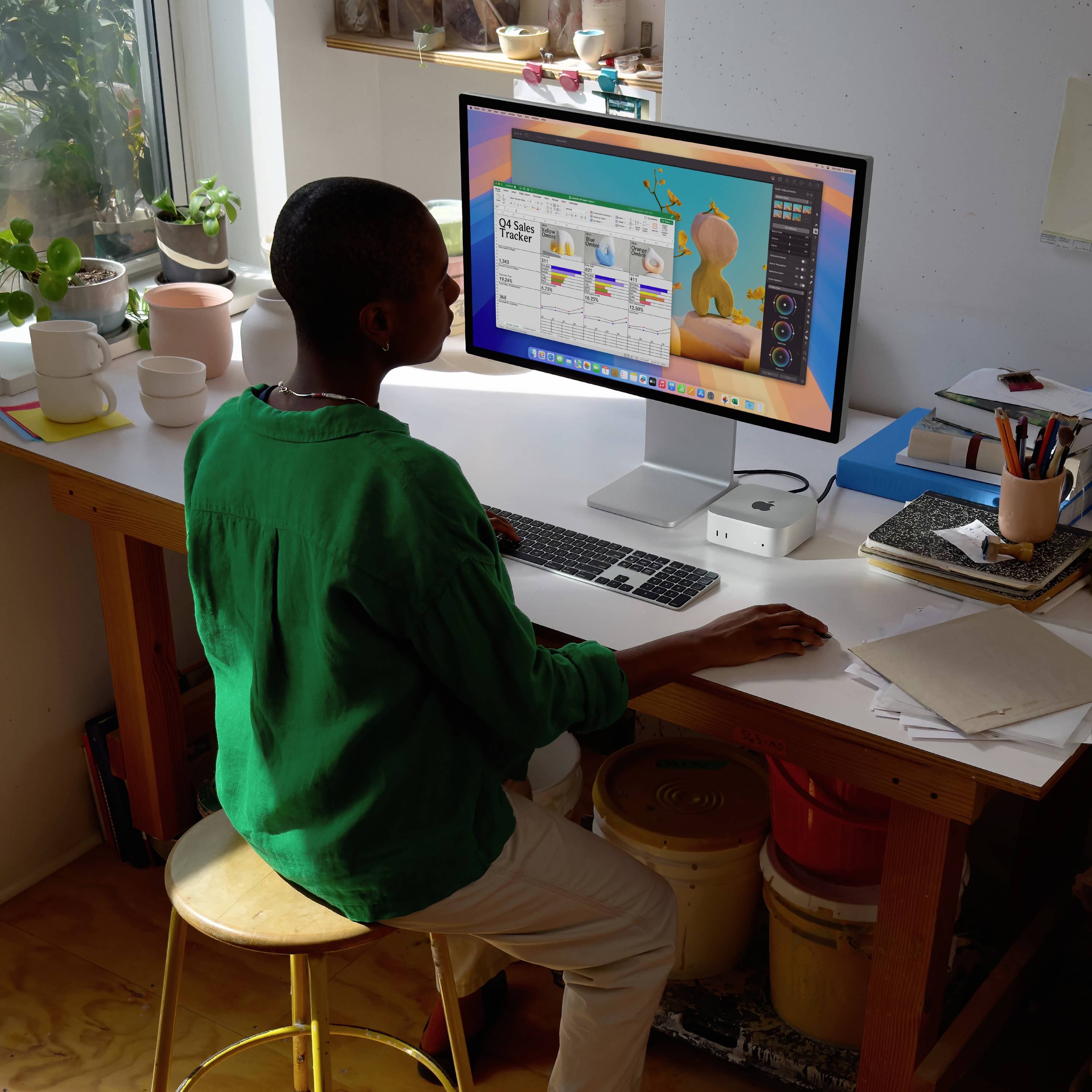 Une personne travaille à un bureau sur un ordinateur avec un logiciel graphique ; un écran lumineux ; des pots et des plantes sur le rebord de la fenêtre.