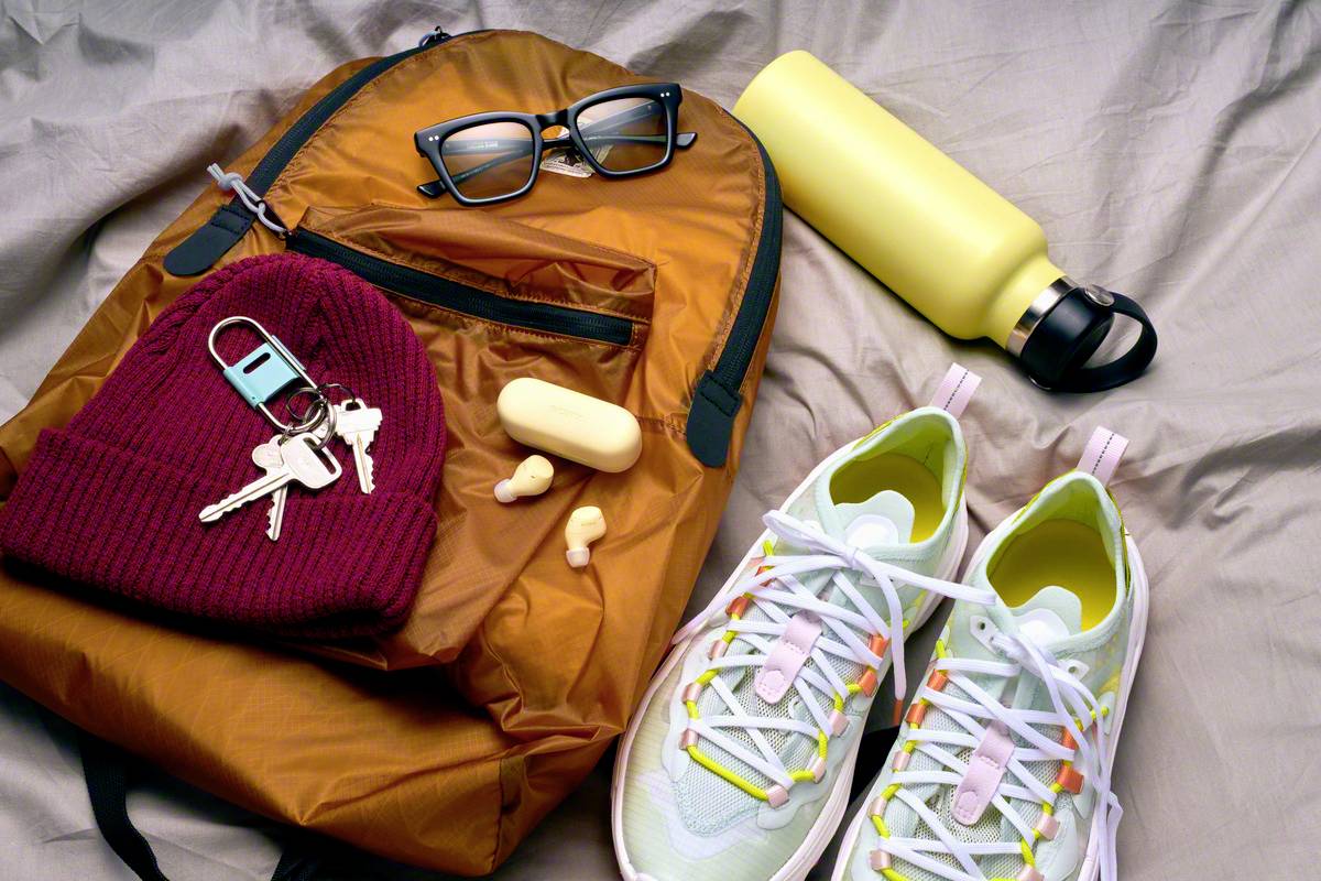 Un sac à dos orange, des lunettes, une gourde jaune, une casquette rouge avec des clés, des écouteurs sans fil et des baskets claires.