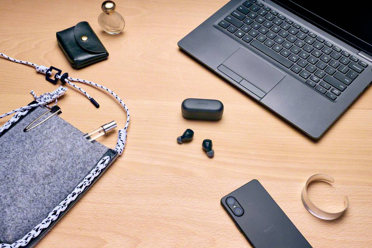 Sur un bureau, un ordinateur portable ouvert est posé, à côté d'un smartphone, d'écouteurs sans fil, d'un carnet de notes, d'un portefeuille et d'un flacon de parfum.