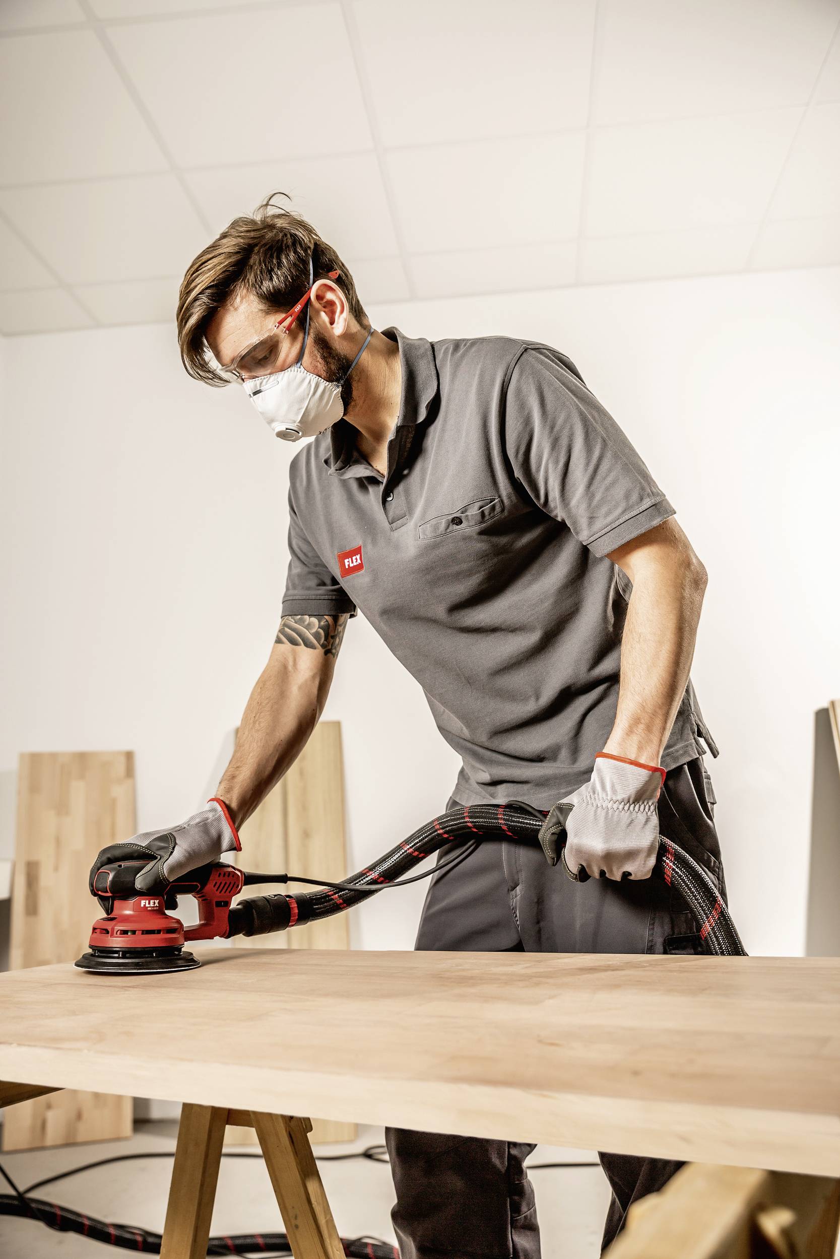 Un homme ponce une planche en bois à l'aide d'une ponceuse électrique ; il porte des lunettes de protection, un masque et des gants dans un atelier.